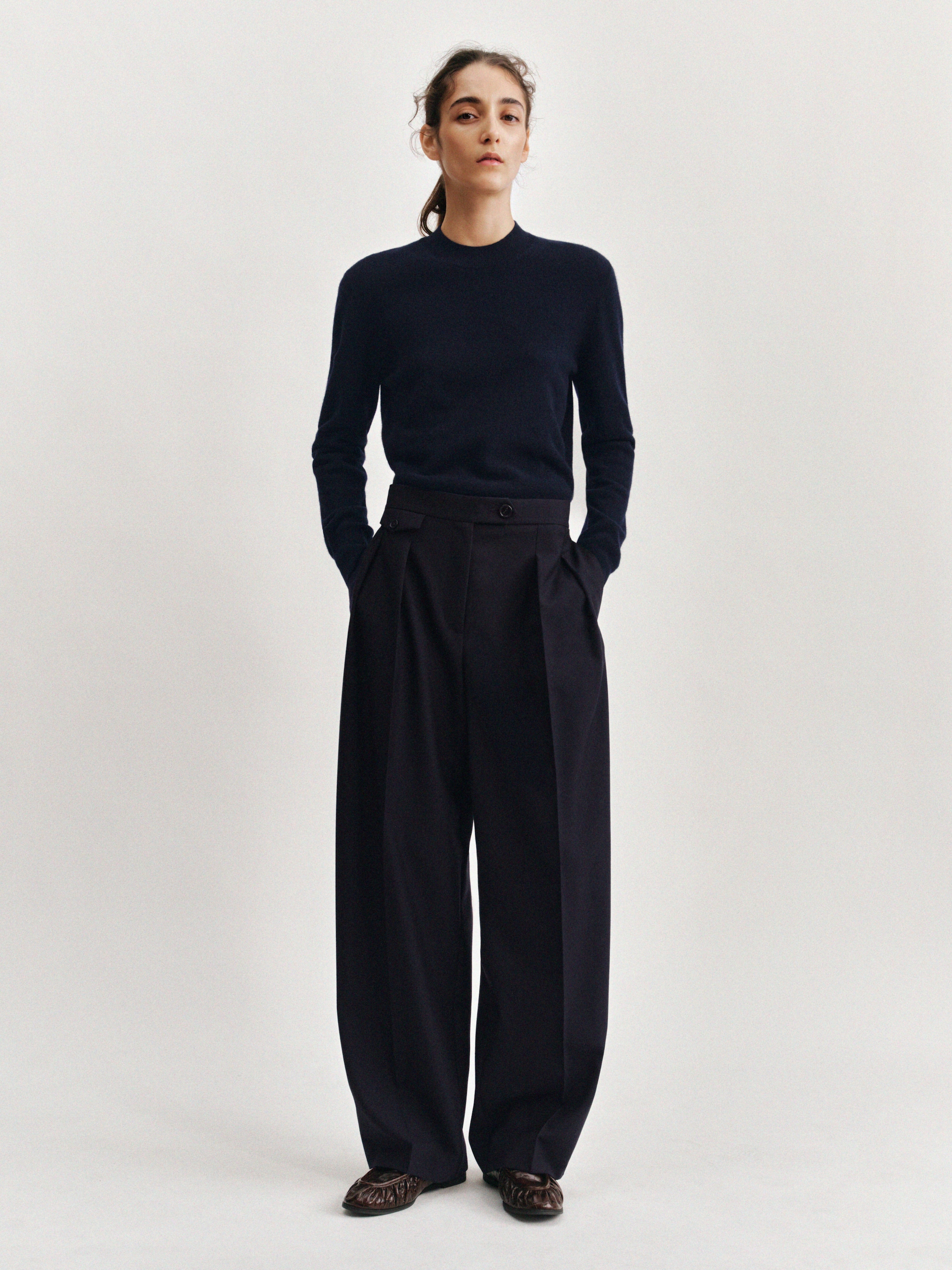 A person stands against a plain background, wearing a dark sweater and wide-leg trousers, with hands in pockets and a neutral expression.