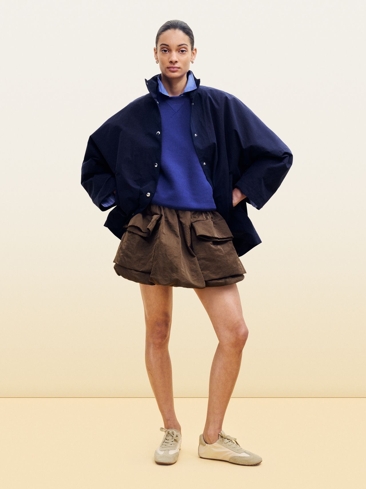 Person wearing an oversized navy jacket, blue shirt, brown skirt, and beige shoes stands confidently against a plain background.