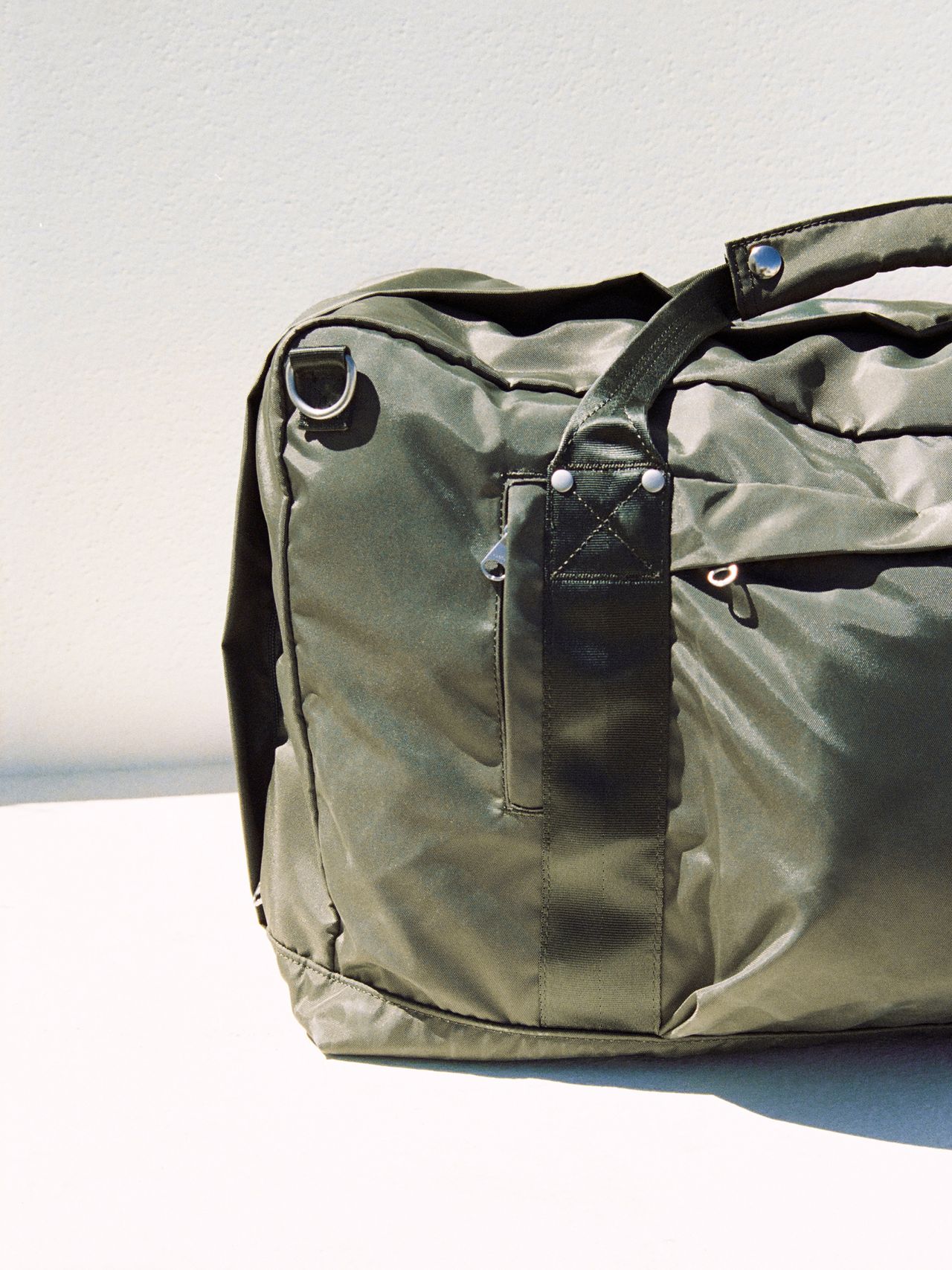 Olive green duffel bag with black handles and metal rings, sitting on a white surface against a light background.