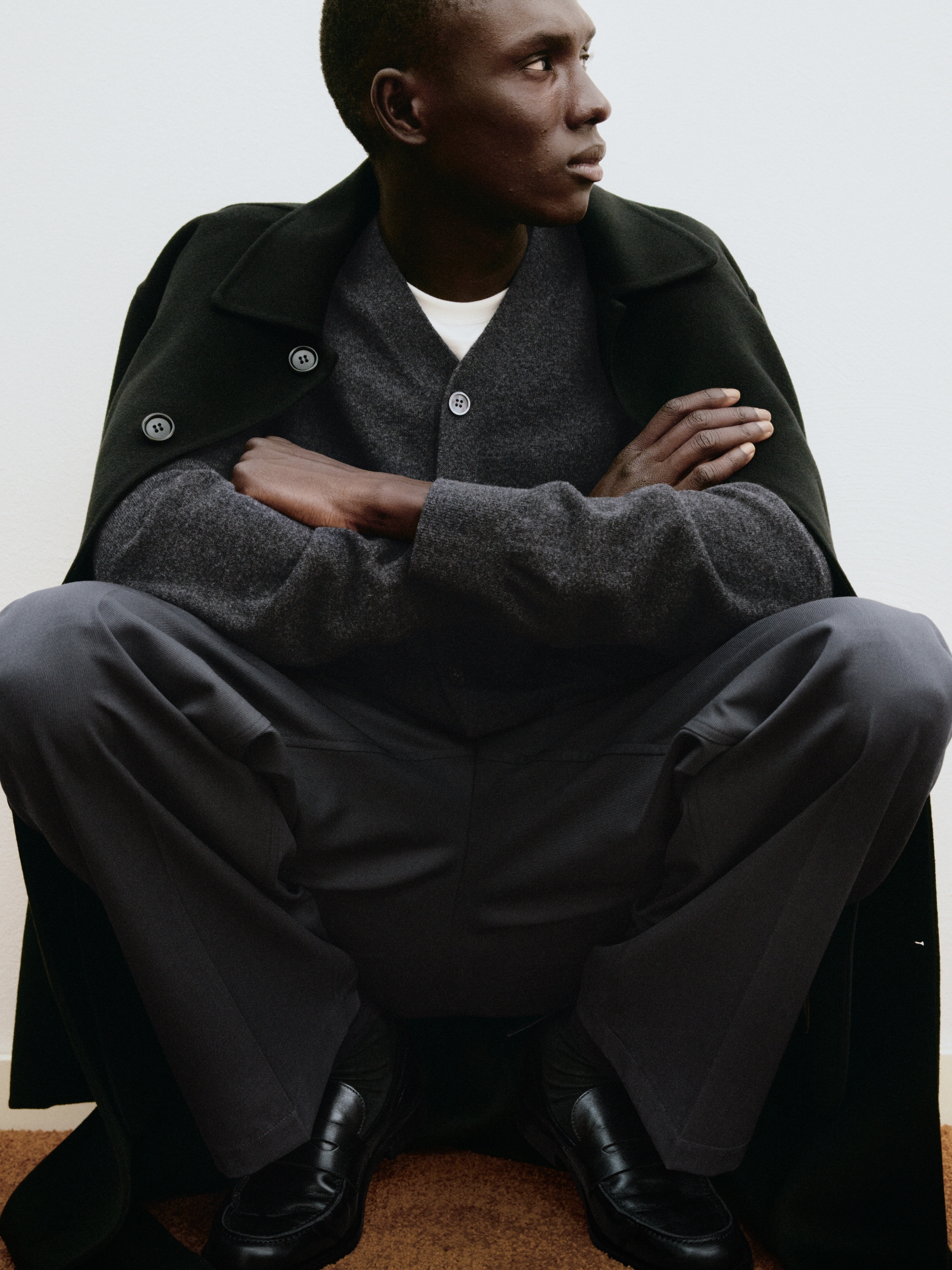 Man in a dark coat and gray sweater sits with crossed arms, looking to the side.
