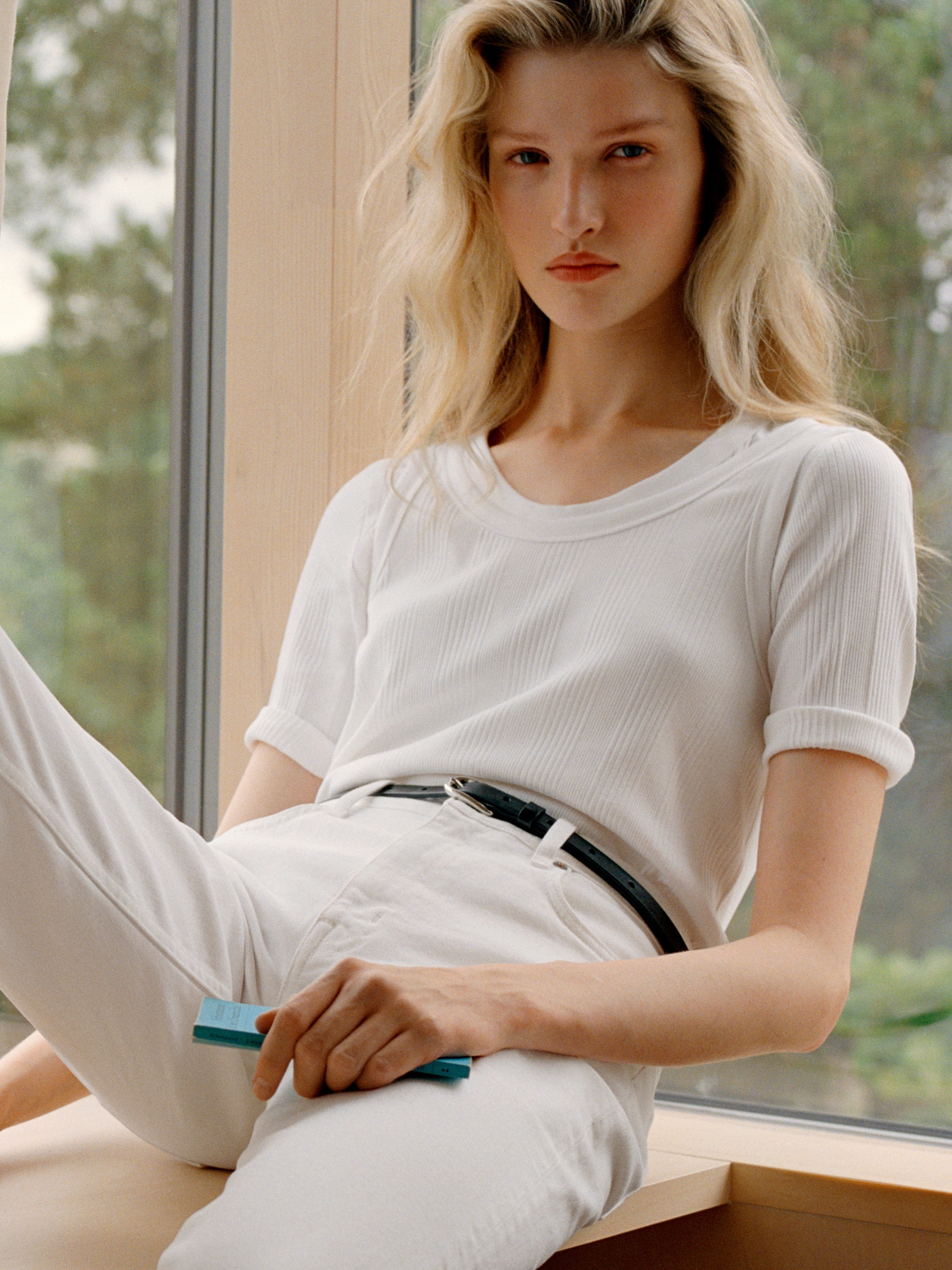 A person with long blonde hair in a white outfit, holding a small blue object, poses by a large window with greenery outside.