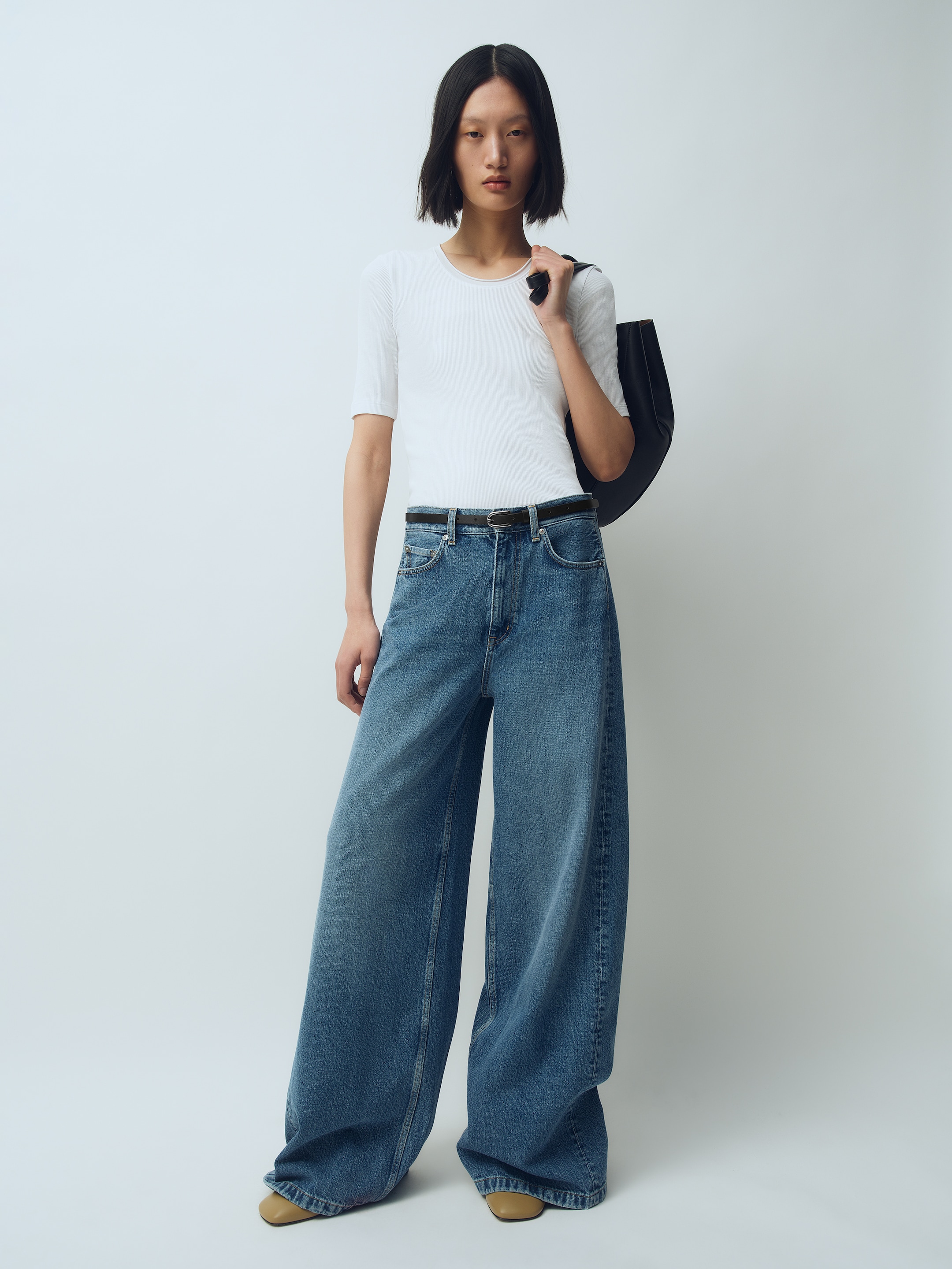 A person with a short hairstyle wears a white top and high-waisted wide-leg jeans, holding a black bag over their shoulder against a plain background.