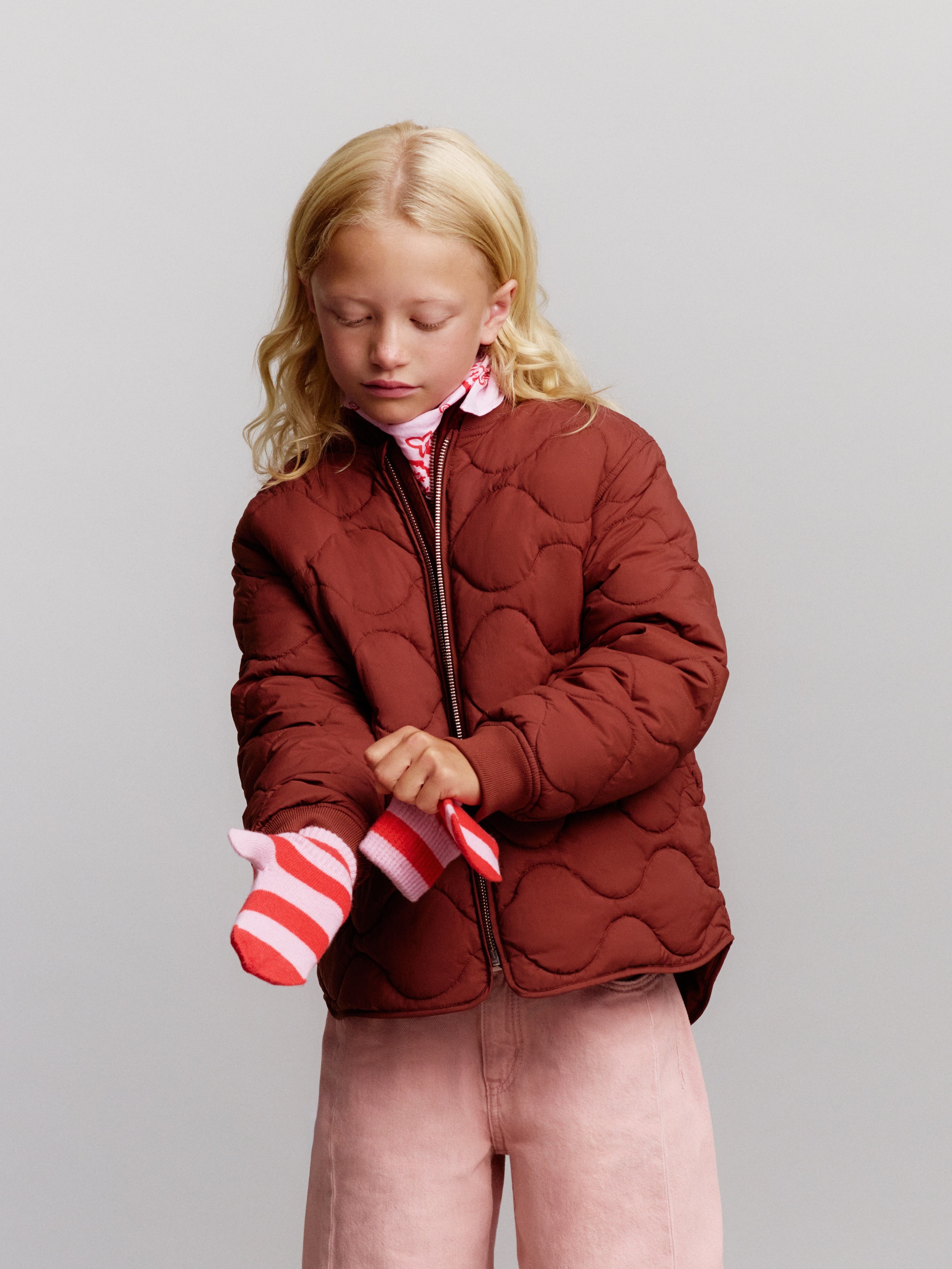 A child with blonde hair wears a brown quilted jacket and pink pants, adjusting a striped red and pink sock.