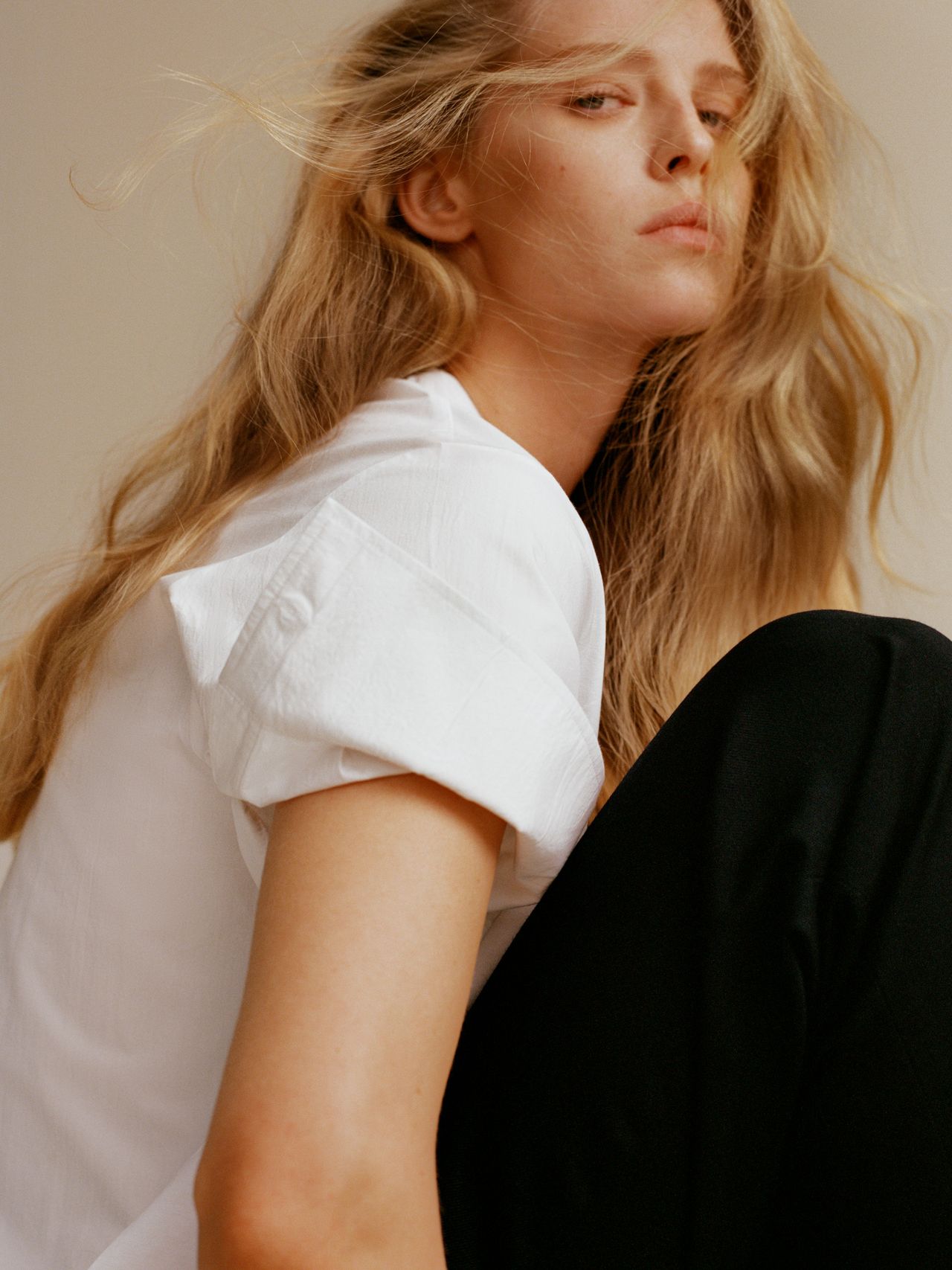 Person with long blonde hair wearing a white shirt and black pants, sitting with a neutral expression, looking slightly upward.
