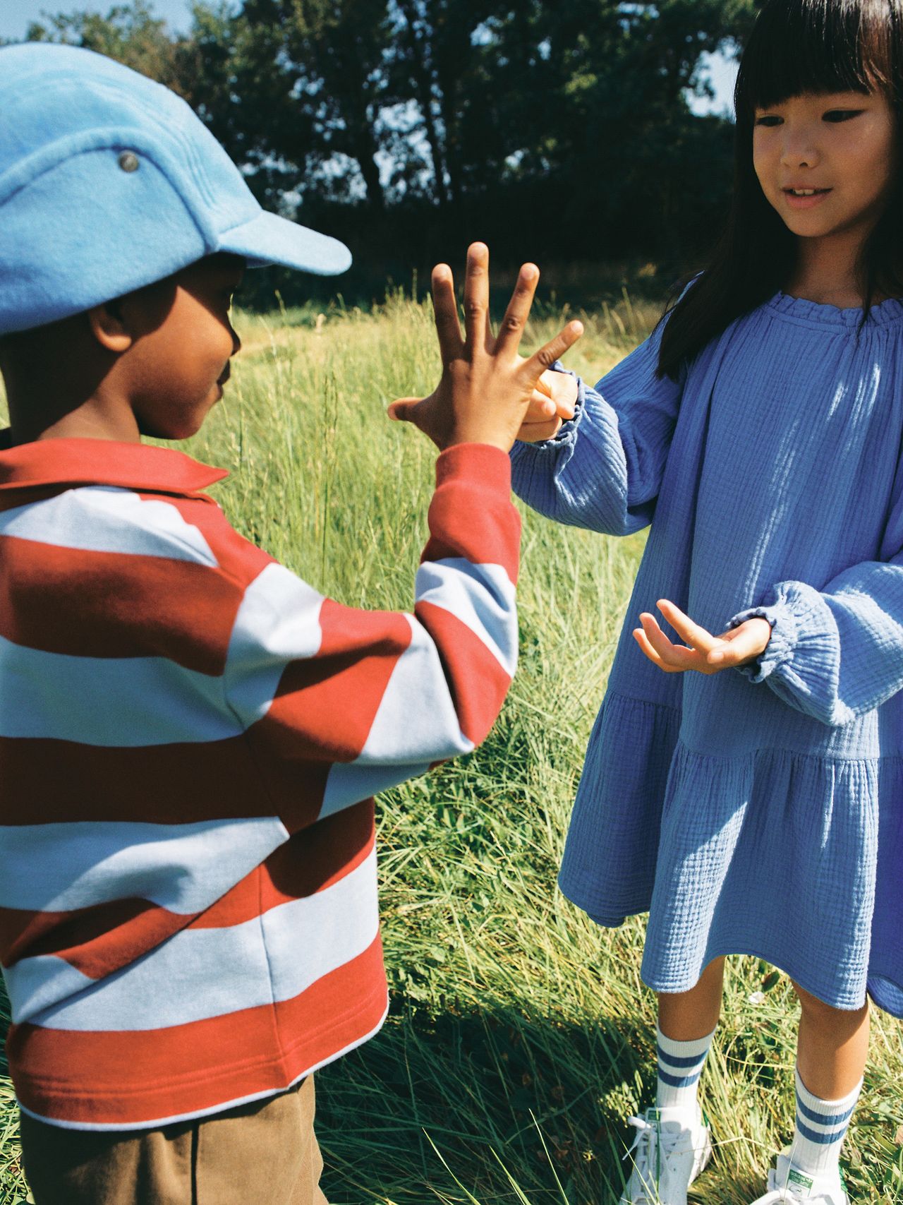 Two children play a hand-clapping game in a grassy field. One wears a striped shirt and blue cap, the other a blue dress. Trees are in the background.