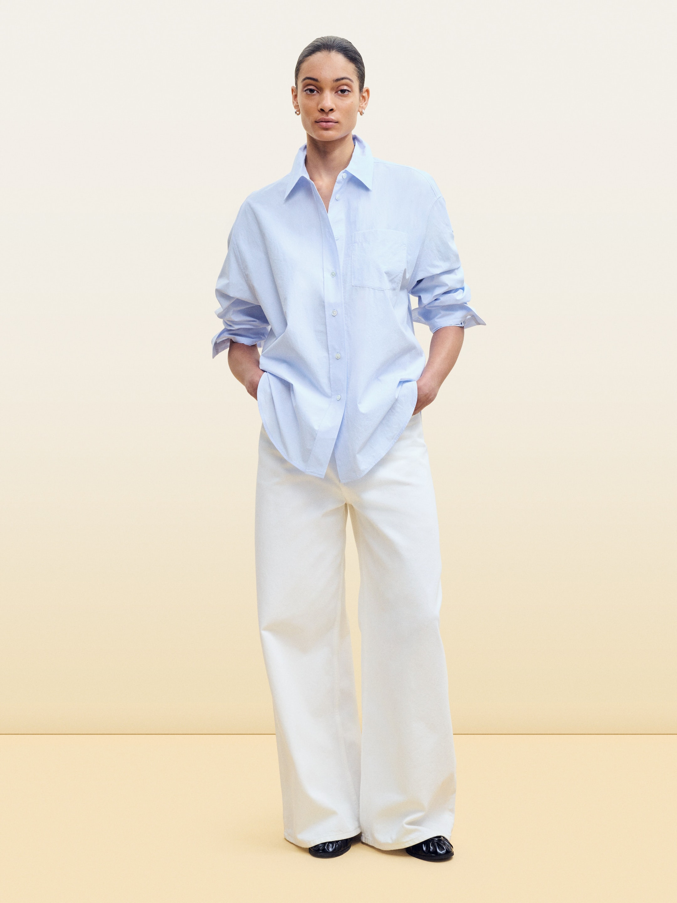 Person in a light blue button-up shirt and white wide-leg pants stands against a plain background, with hands in pockets.