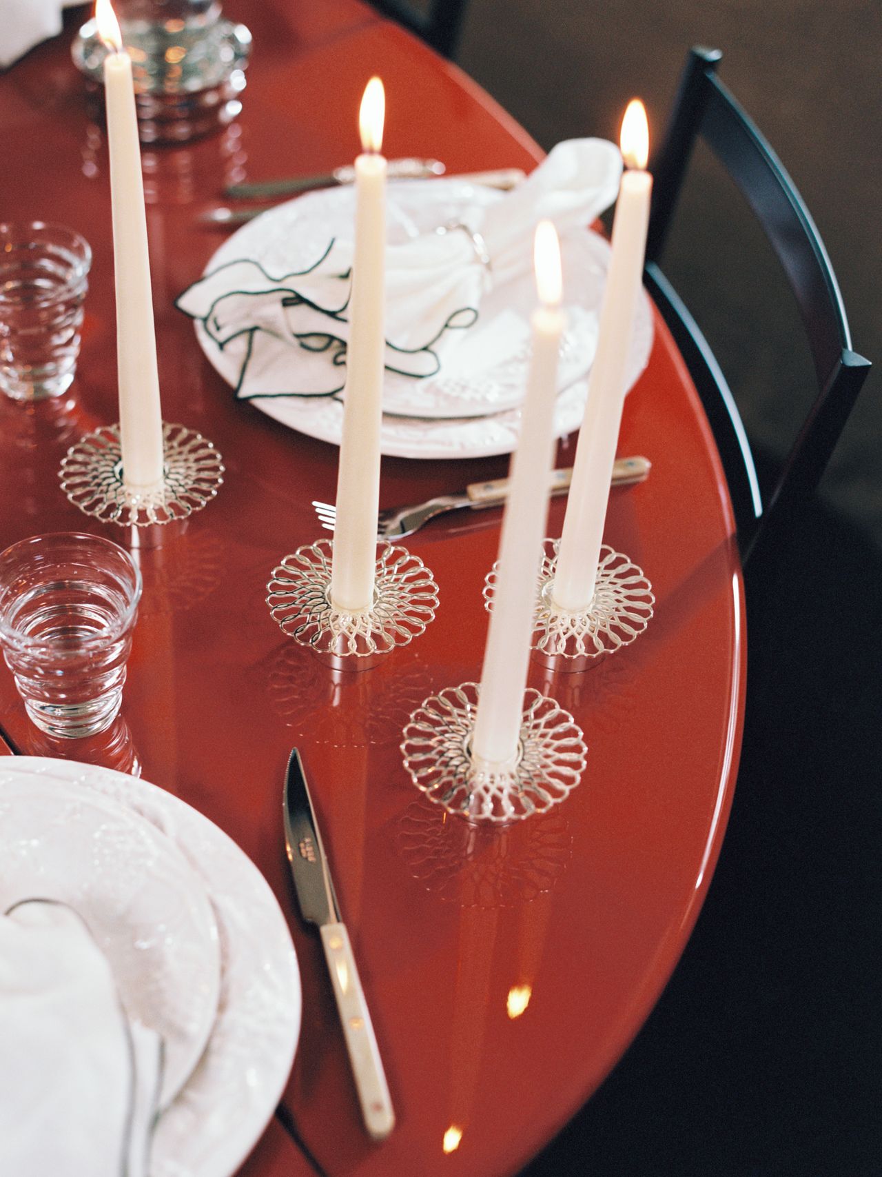 A red table set for a meal with white plates, silverware, glasses, and three lit white candles in ornate holders.
