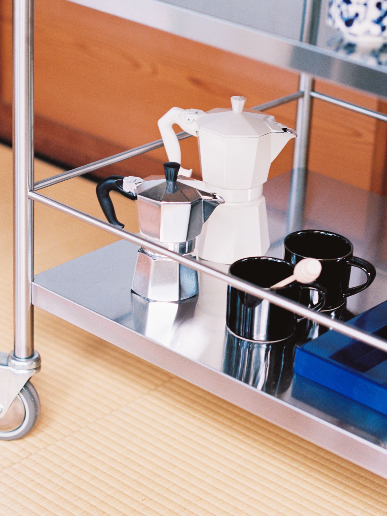A stainless steel cart with a white and a silver coffee maker, black mugs, and a blue box on a woven mat floor.