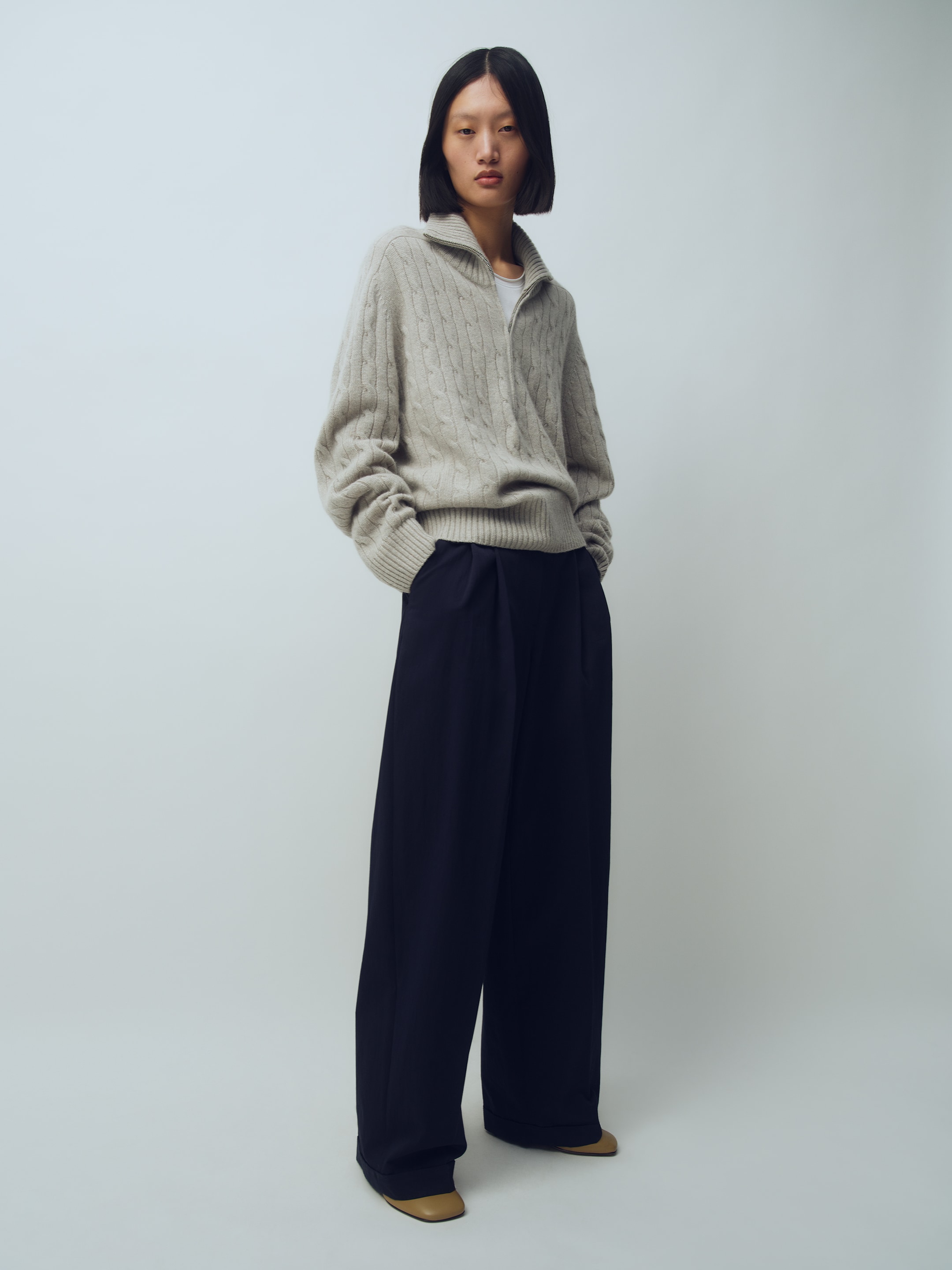 Person with short hair wearing a gray cable-knit sweater, wide-leg black pants, and brown shoes, standing against a plain background.