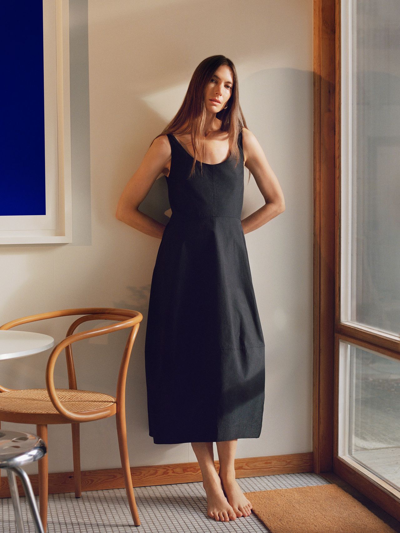 A woman in a sleeveless black dress stands barefoot in a sunlit room with wooden chair and table, casting soft shadows on the wall.