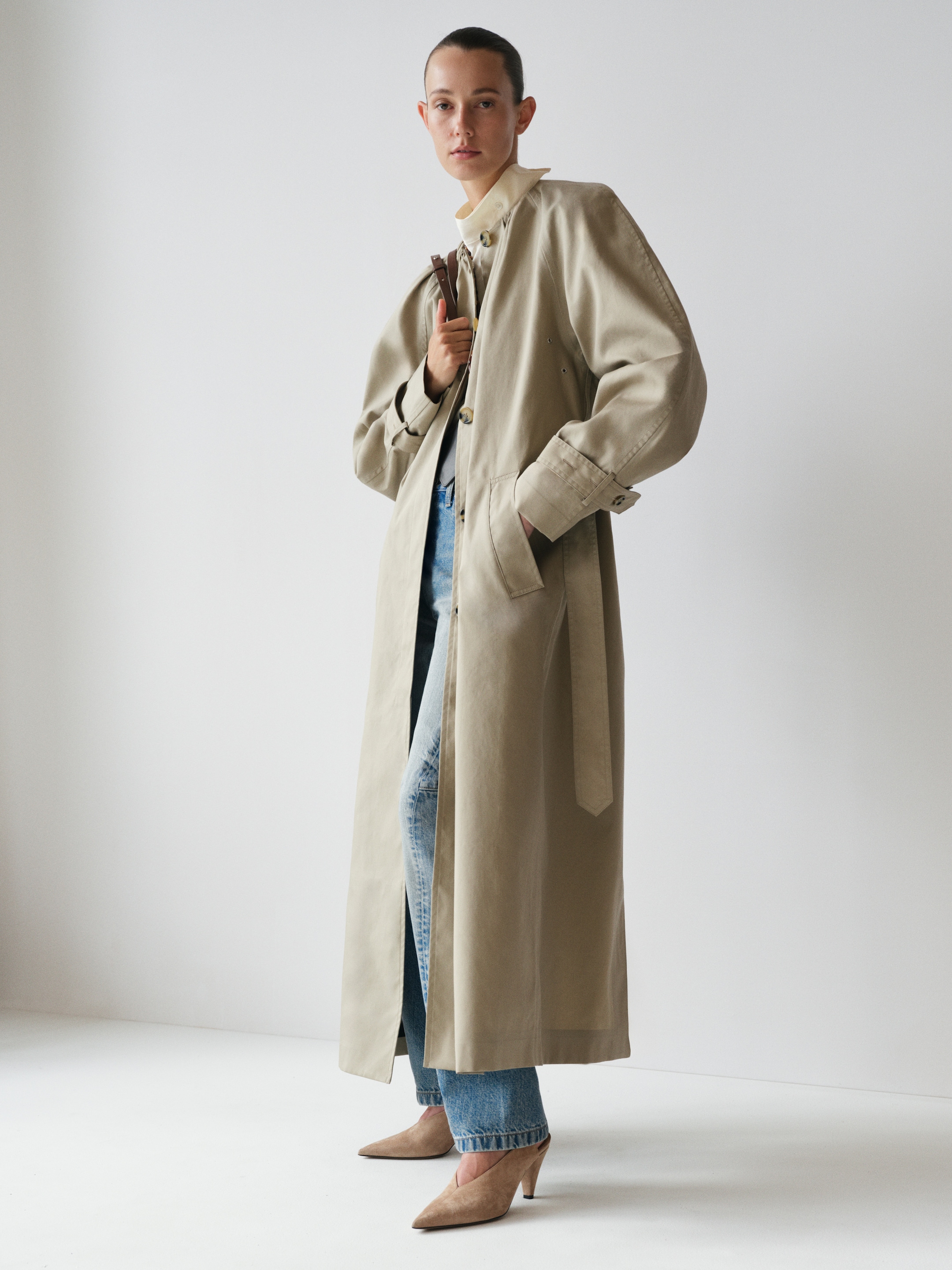 Person in a long beige trench coat, blue jeans, and tan heels, standing against a plain white background, holding the coat collar.