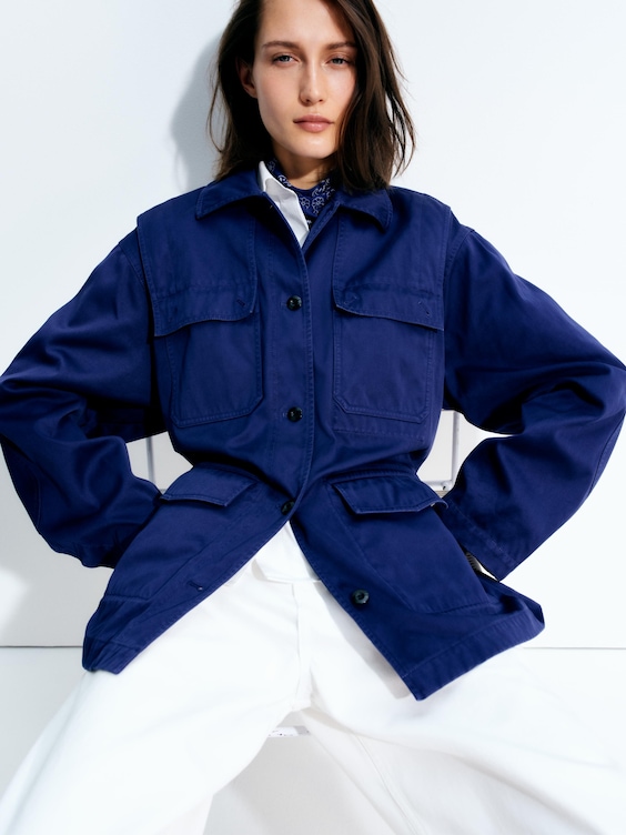 Person wearing a blue utility jacket over a white outfit, sitting against a white background with a confident posture.
