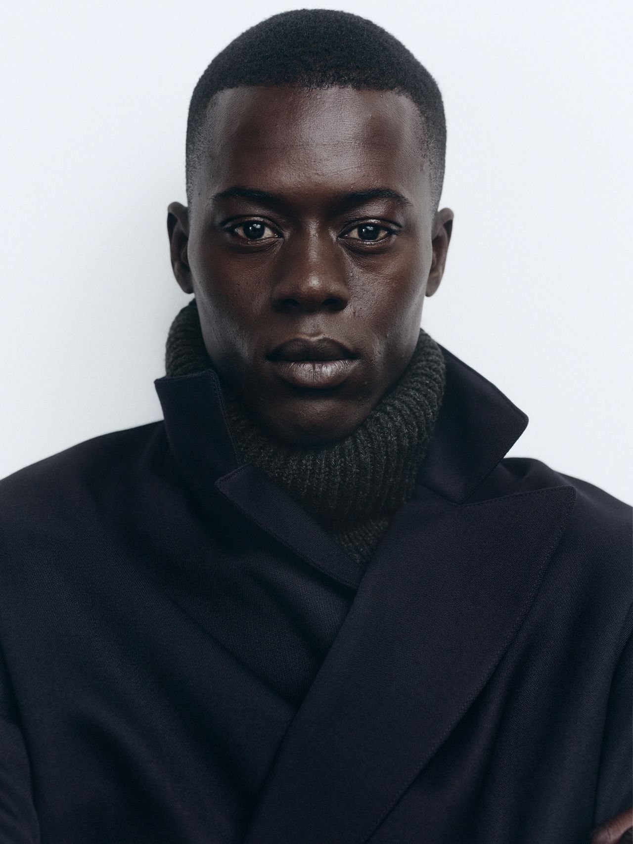 Person wearing a dark coat and a knitted scarf, looking directly at the camera against a plain background.