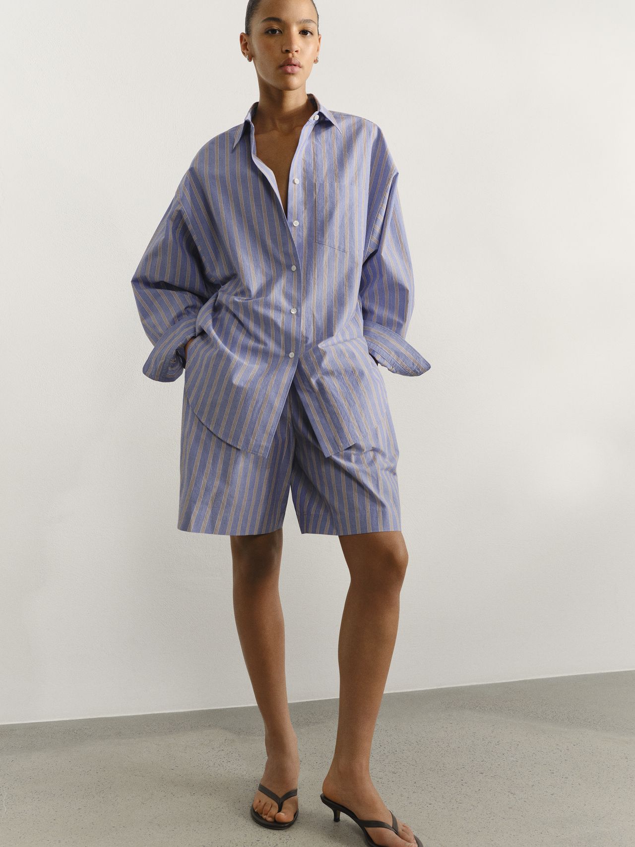 Person wearing a blue and white striped oversized shirt and matching shorts with black sandals, standing against a plain background.
