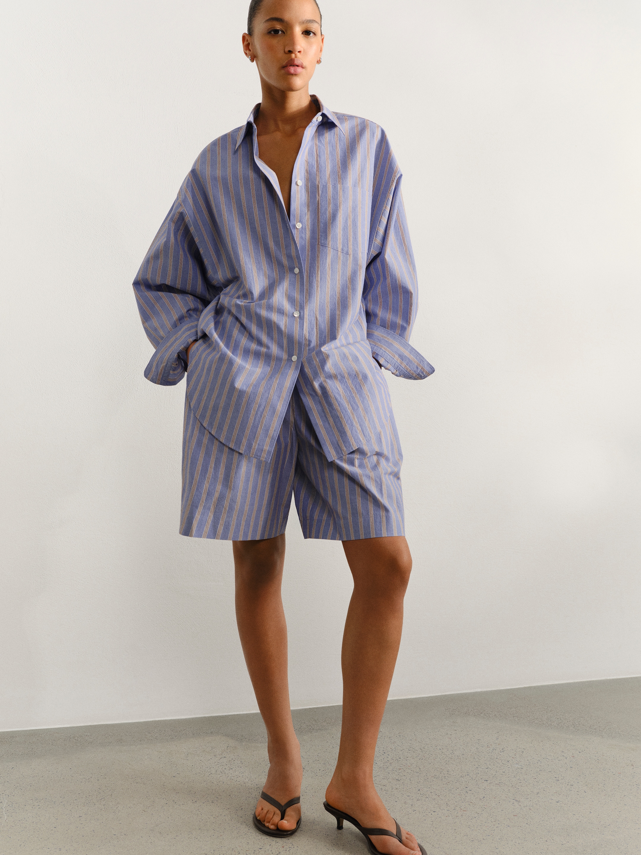 Person wearing a blue and white striped oversized shirt and matching shorts with black sandals, standing against a plain background.