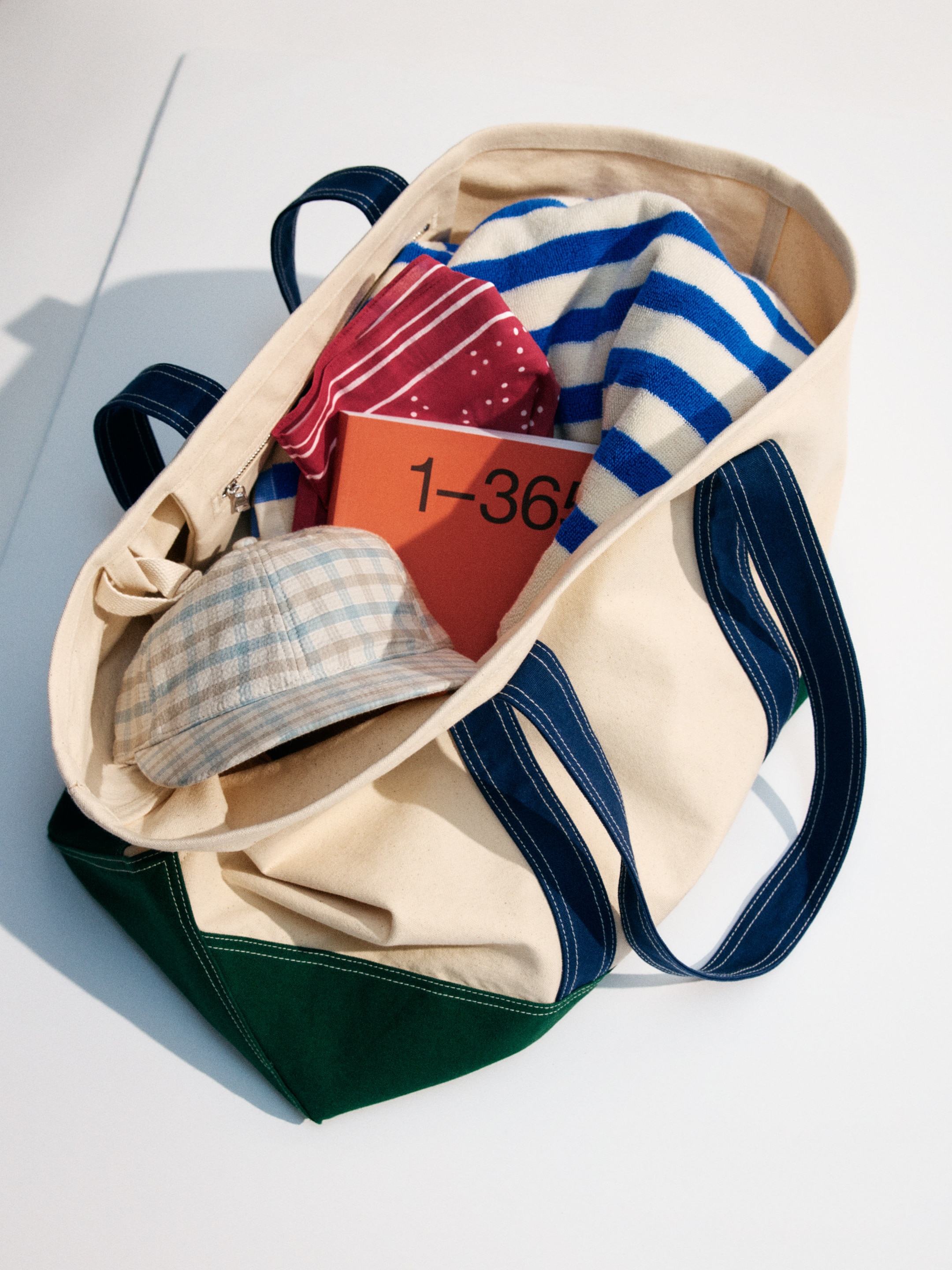 Open canvas tote bag with blue handles, holding a blue-striped towel, red scarf, cap, and an orange book labeled "1-365."