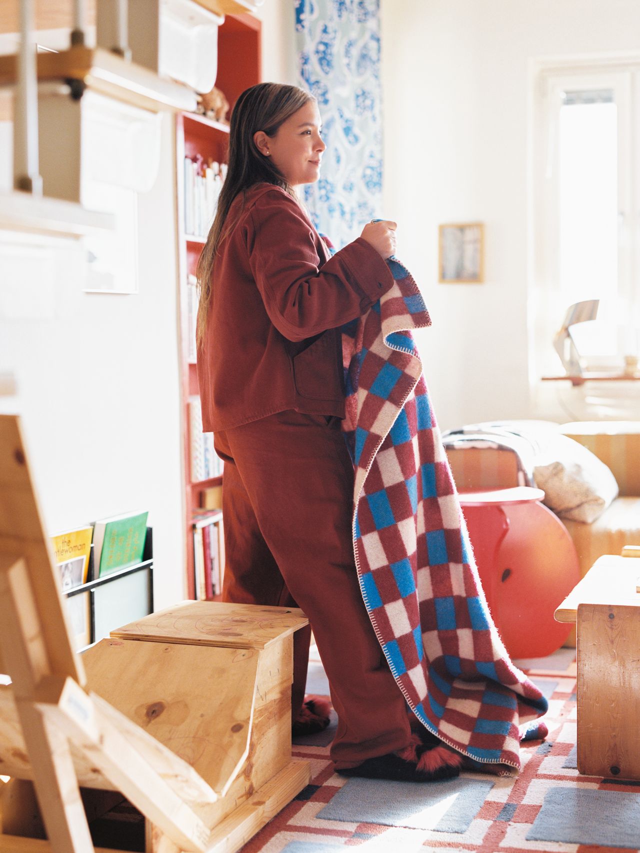 A person in a cozy room holds a colorful checkered blanket, standing near wooden furniture and shelves filled with books.