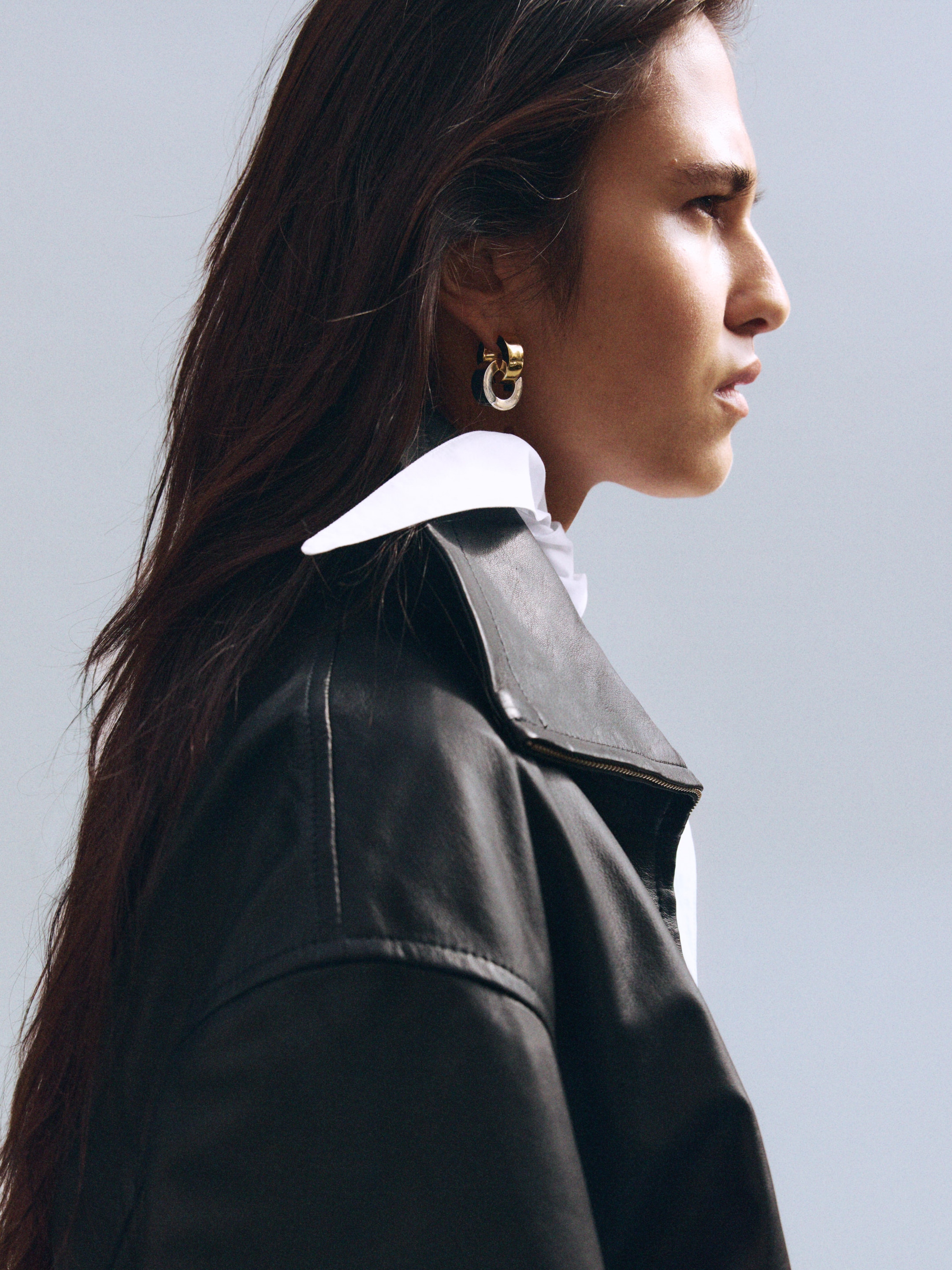Person in profile with long hair, wearing a black leather jacket and gold hoop earrings, set against a neutral background.