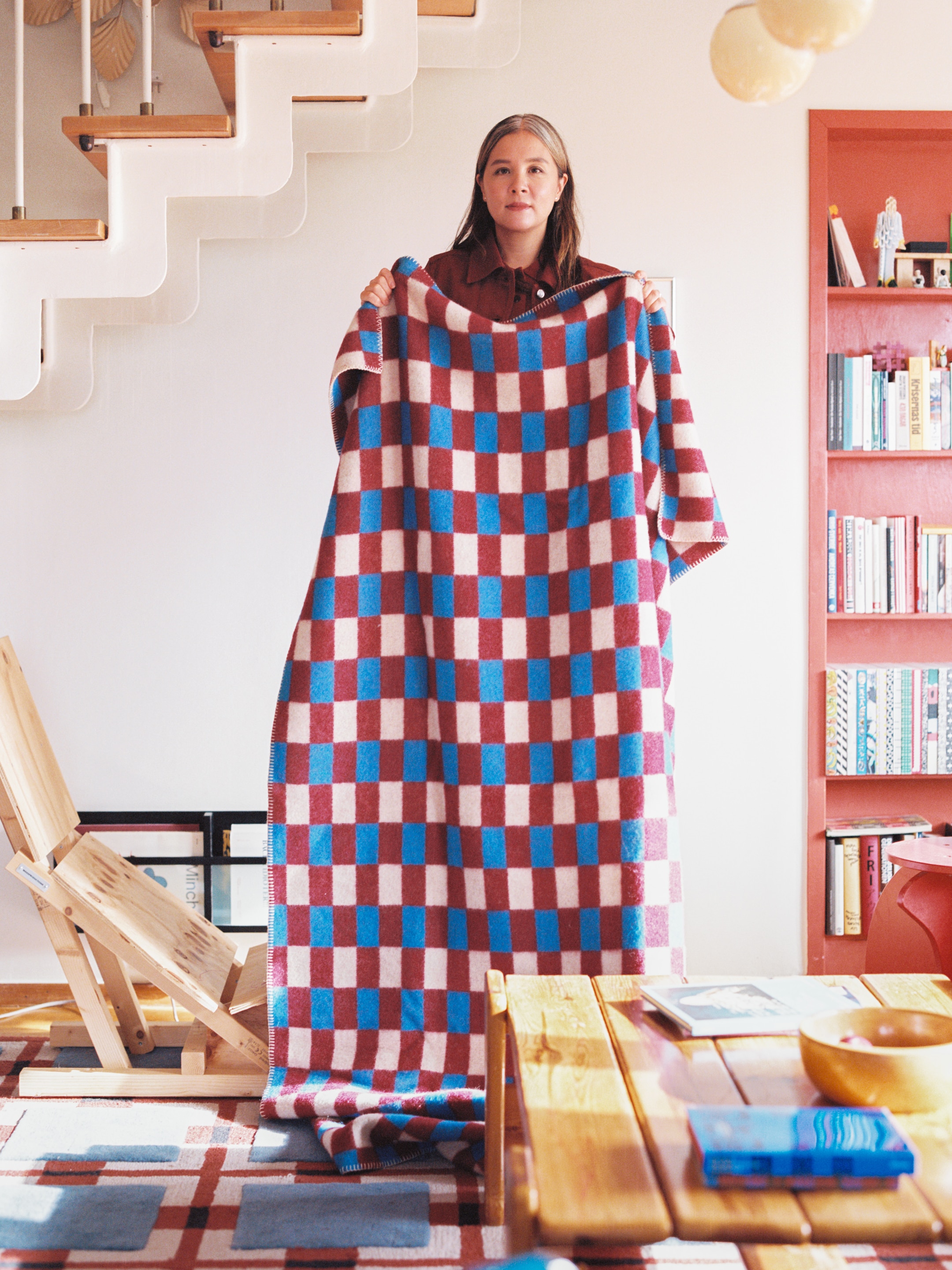 Person holding a large checkered blanket in a cozy room with bookshelves, a wooden table, and a staircase in the background.