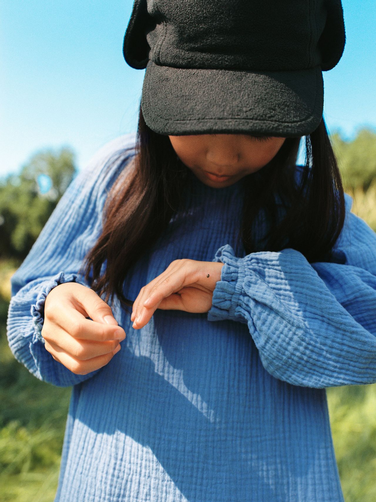 A person in a blue shirt and black cap looks at a ladybug on their hand outdoors, with greenery in the background.