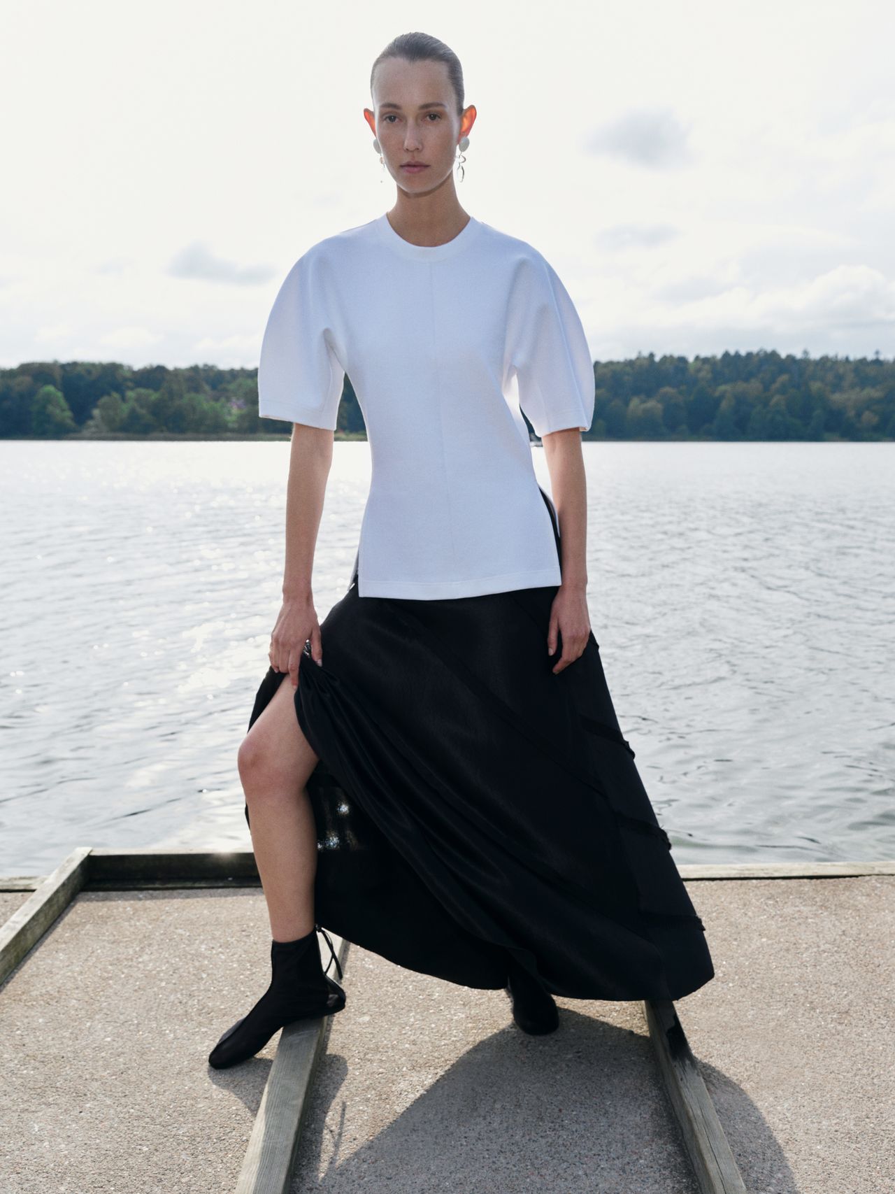 A person stands on a dock by a lake, wearing a white top with puffed sleeves and a long black skirt, with trees in the background.