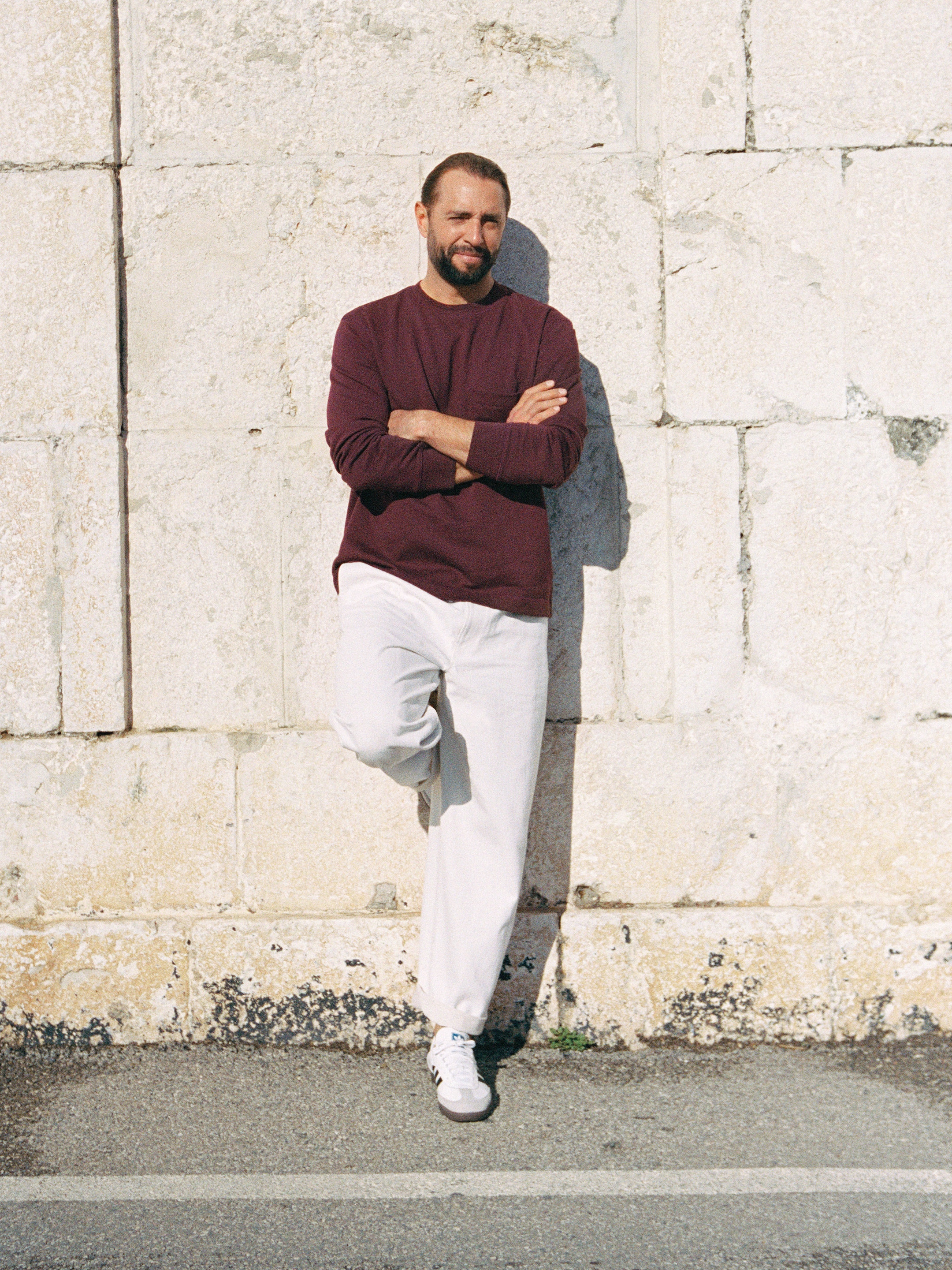 Man in a maroon sweater and white pants leans against a stone wall with arms crossed, one leg bent, and wearing white sneakers.