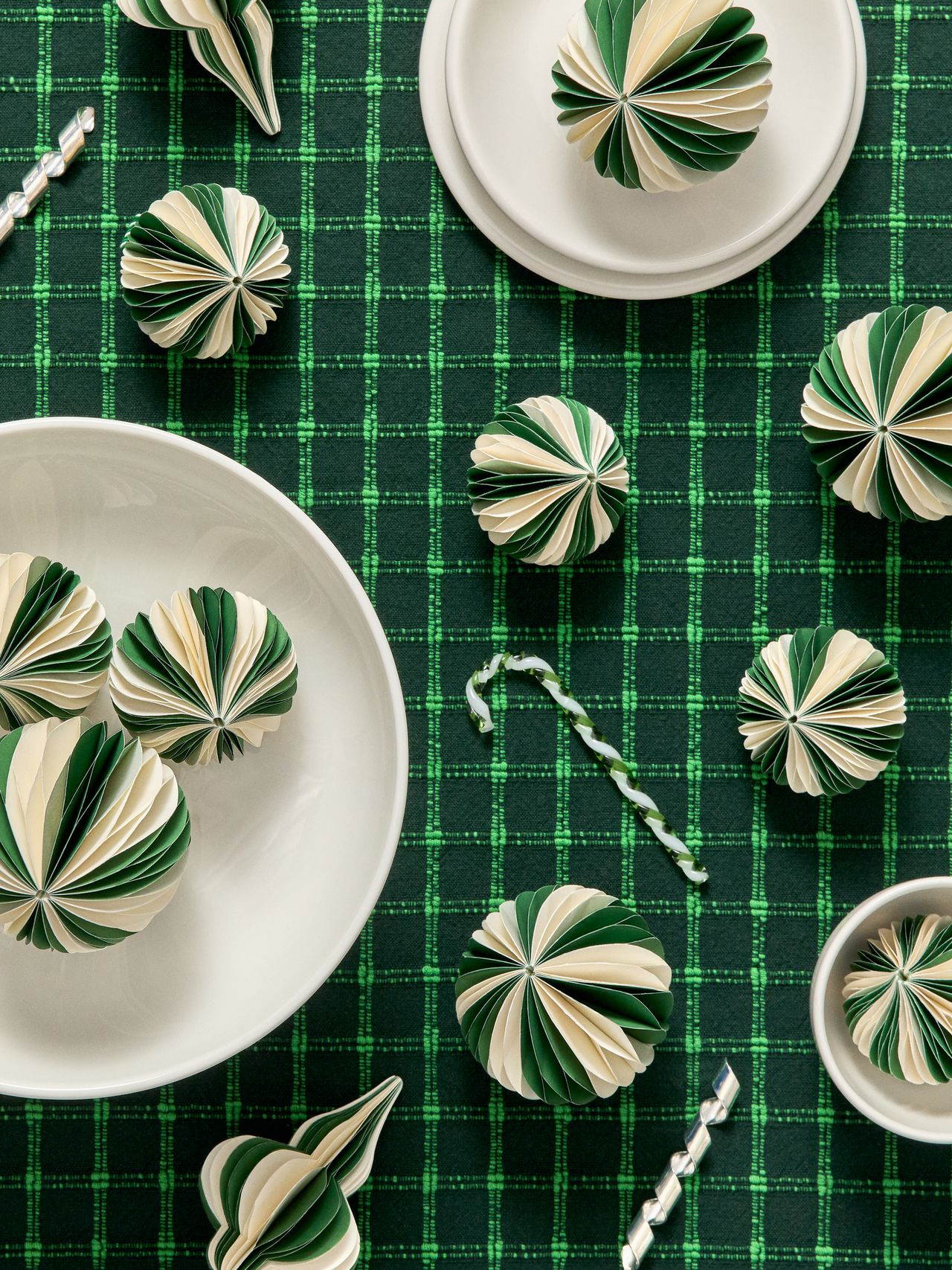Green and white paper ornaments arranged on a green plaid surface with white bowls and silver decorative sticks.