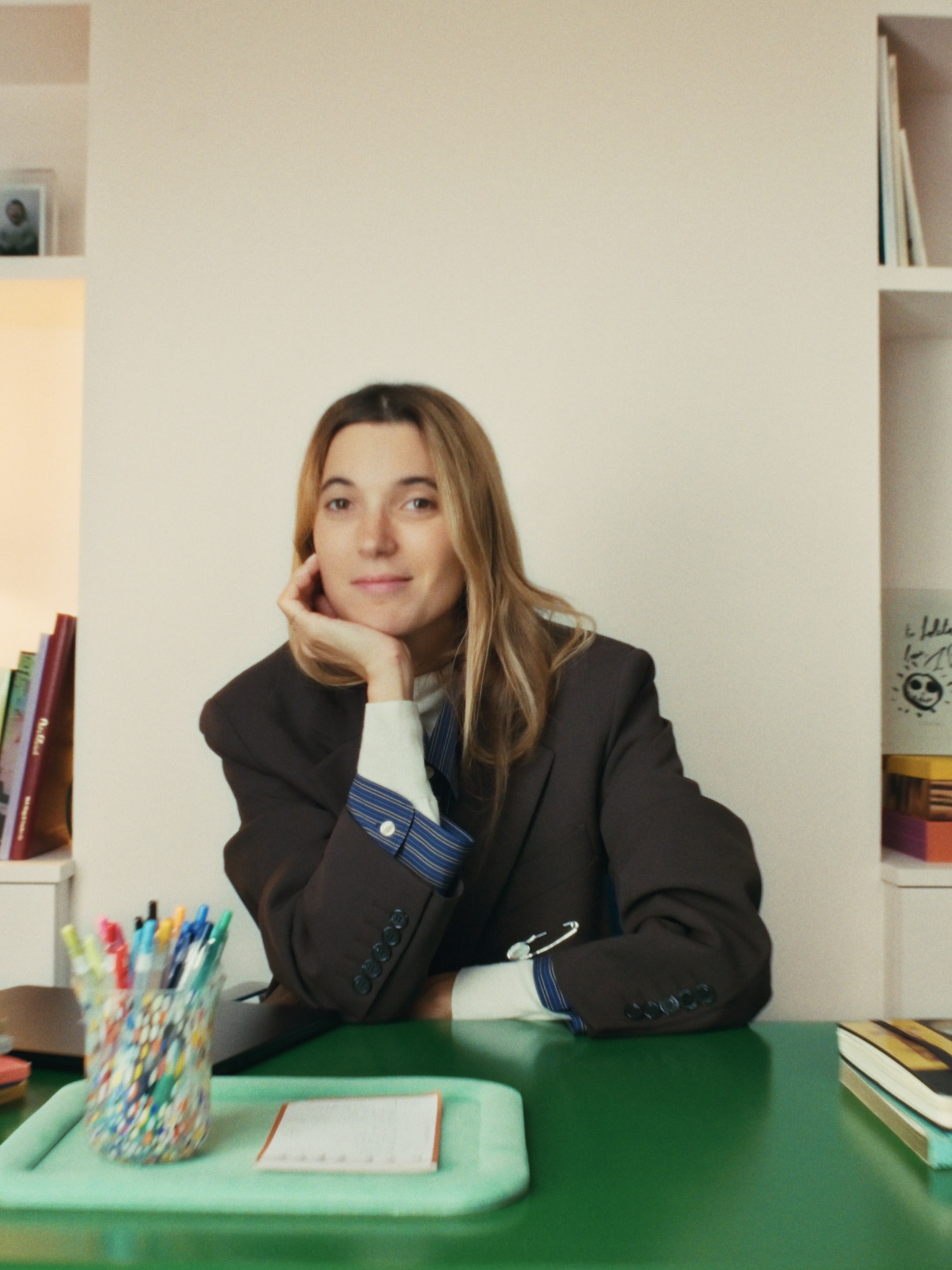 A woman with long hair rests her chin on her hand, sitting at a green desk with colorful pens and books in the background.