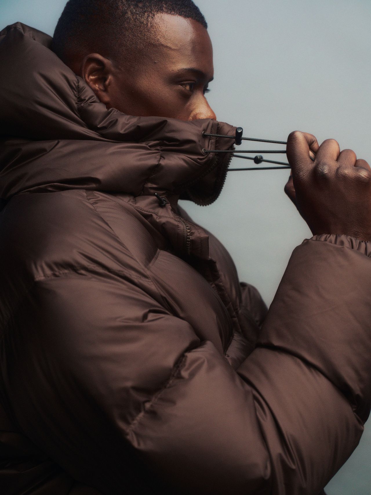 Person in a brown puffer jacket adjusting the hood drawstring, viewed from the side against a neutral background.