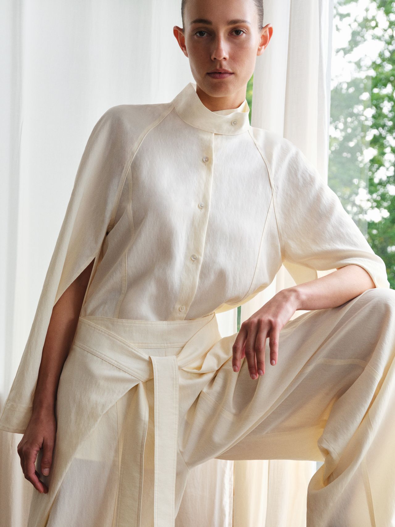 Person in a cream outfit with a high collar and wide-legged pants, posing indoors near sheer curtains with greenery visible outside.