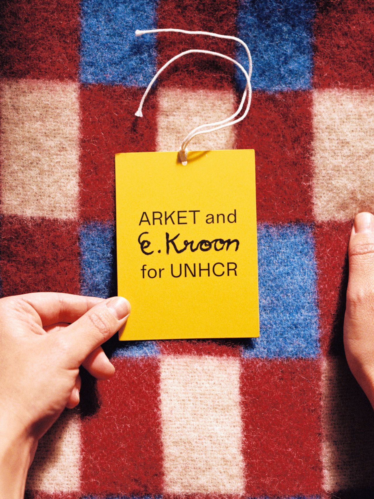 Hands holding a yellow tag reading "ARKET and E. Kroon for UNHCR" on a textured red and blue checkered fabric background.