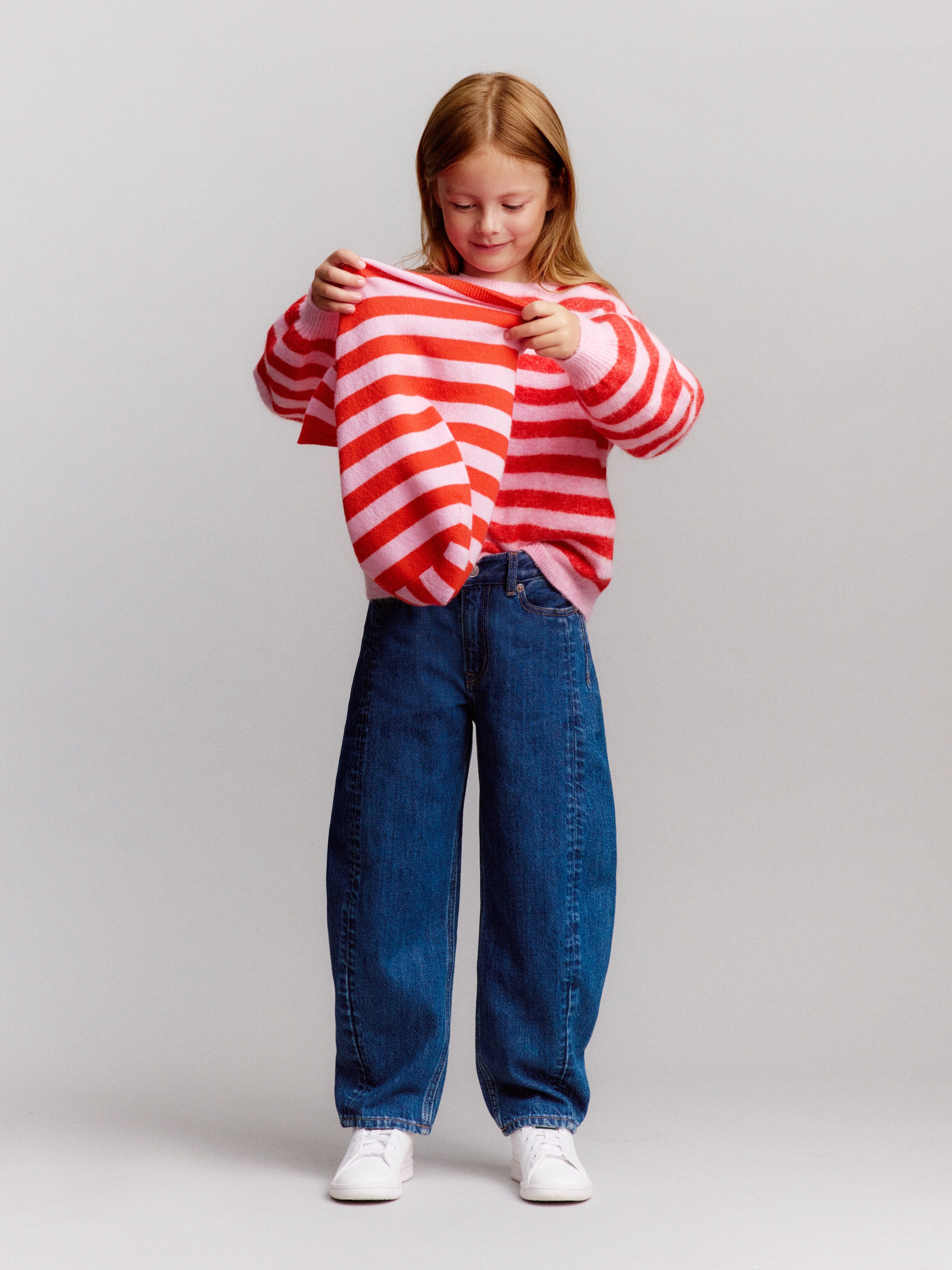 A young girl wearing a red and pink striped sweater and blue jeans looks at her shirt while smiling. She stands against a plain background.