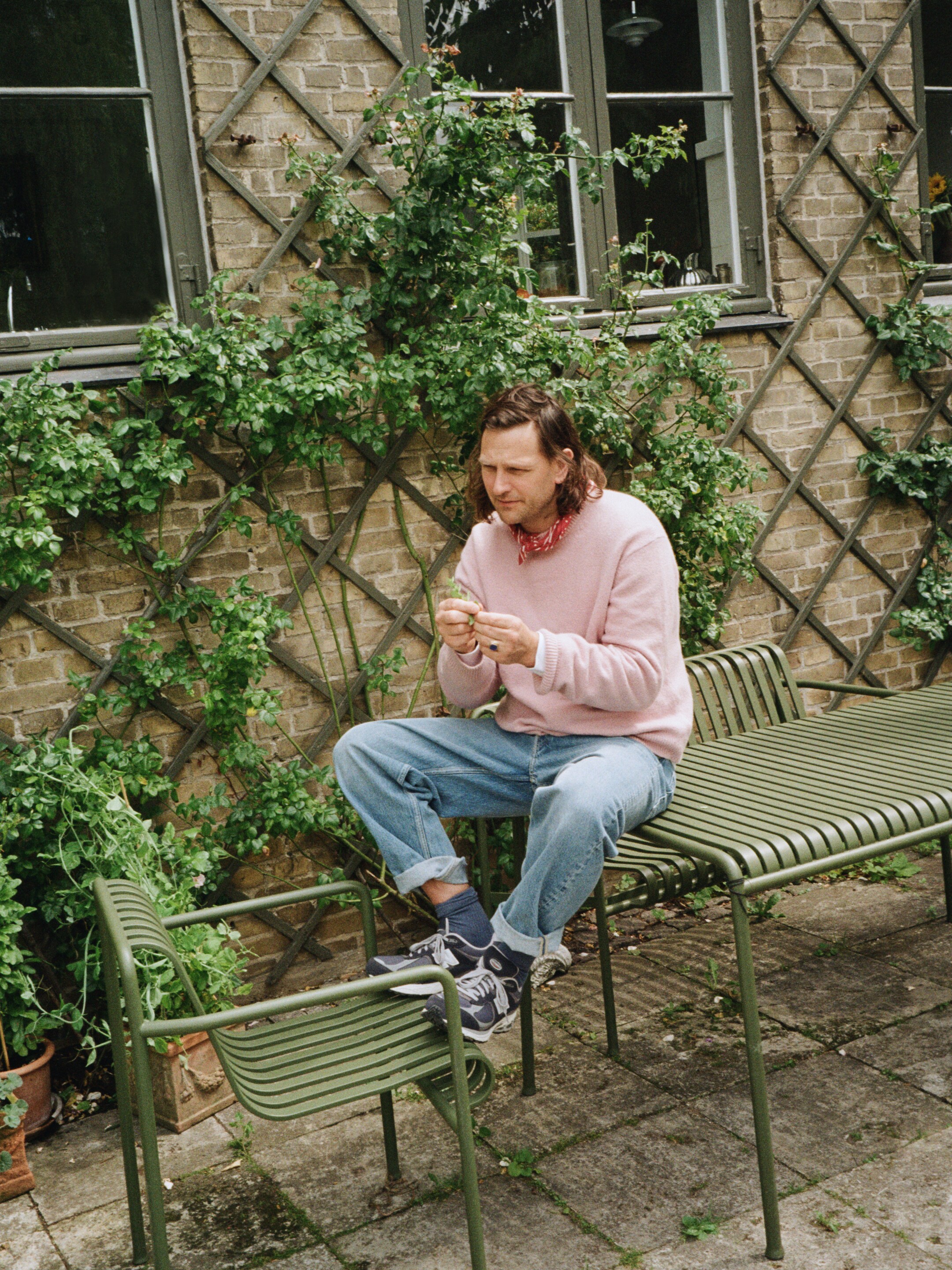 A person in a pink sweater and jeans sits on an outdoor bench, inspecting leaves, with green plants climbing a brick wall behind them.