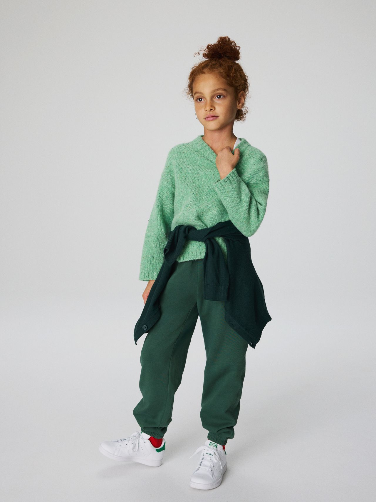 Young girl in a green sweater and pants, with a dark green jacket tied at the waist, stands confidently against a plain background.