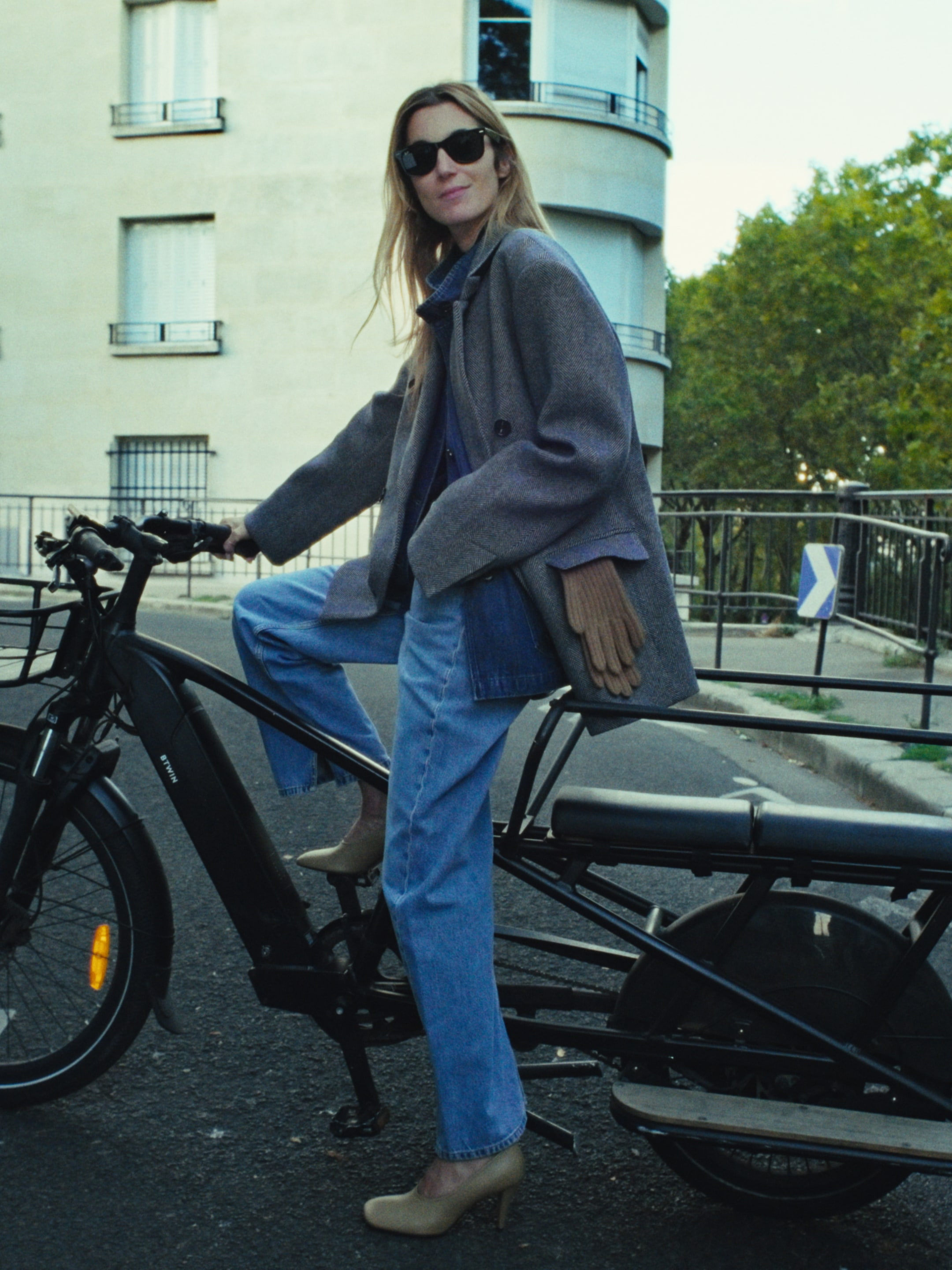 A woman in sunglasses and a coat sits on an e-bike on a city street, her foot on the pedal, with buildings and greenery in the background.