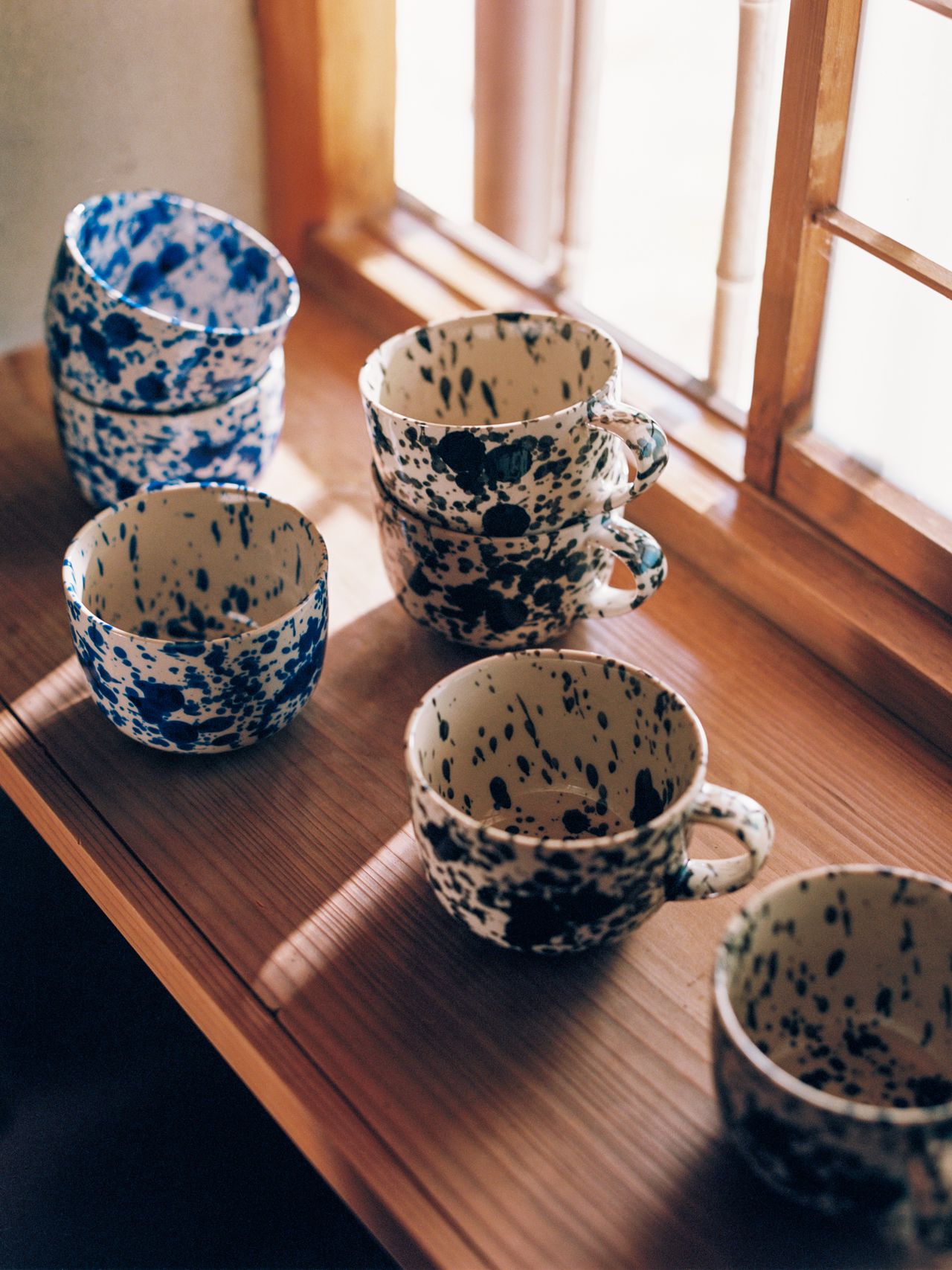 Stacked blue and black speckled ceramic cups and bowls on a wooden windowsill, with sunlight streaming through a window.