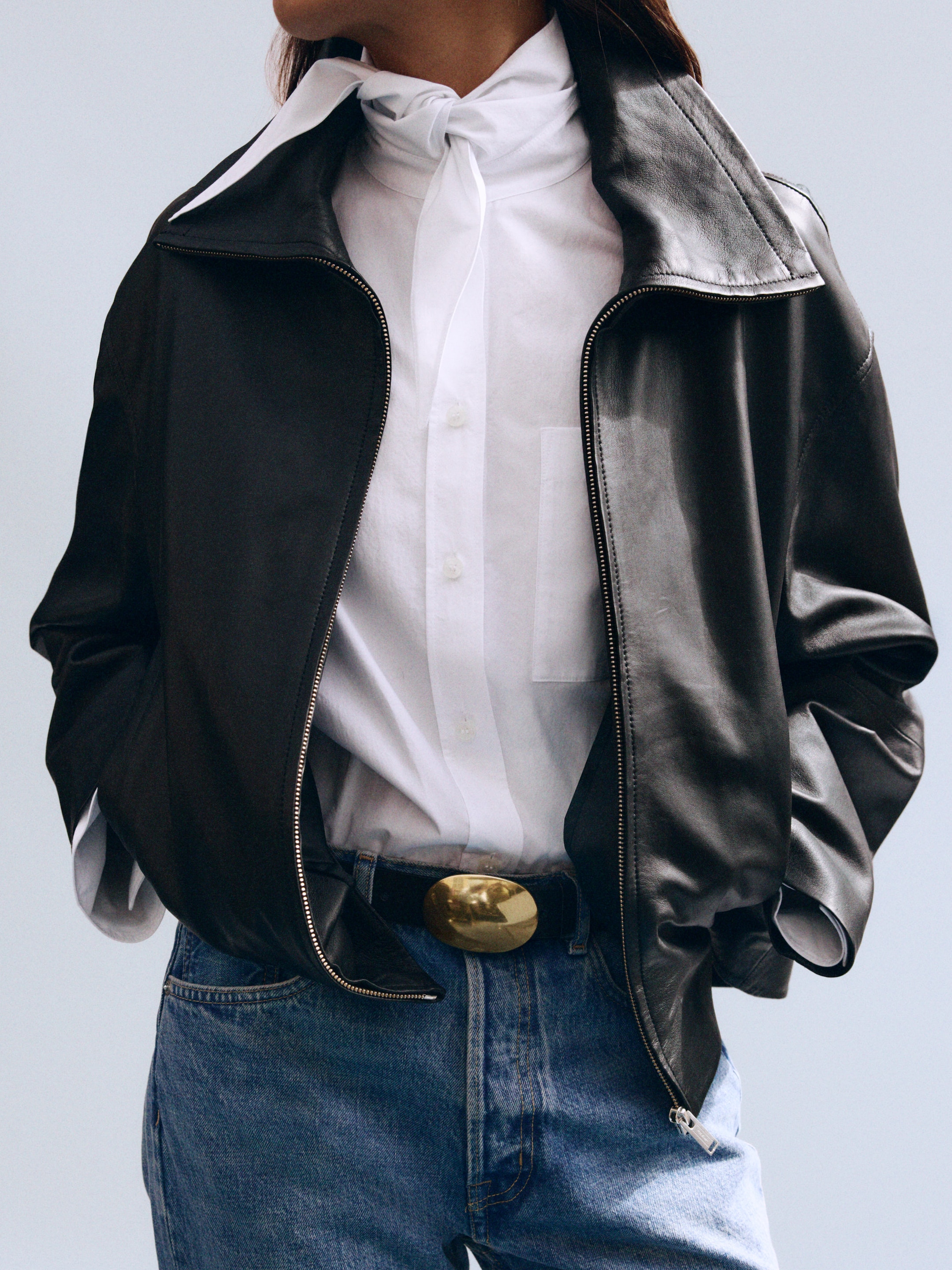 Person wearing a black leather jacket over a white shirt with a neck tie, jeans, and a belt with a gold buckle.