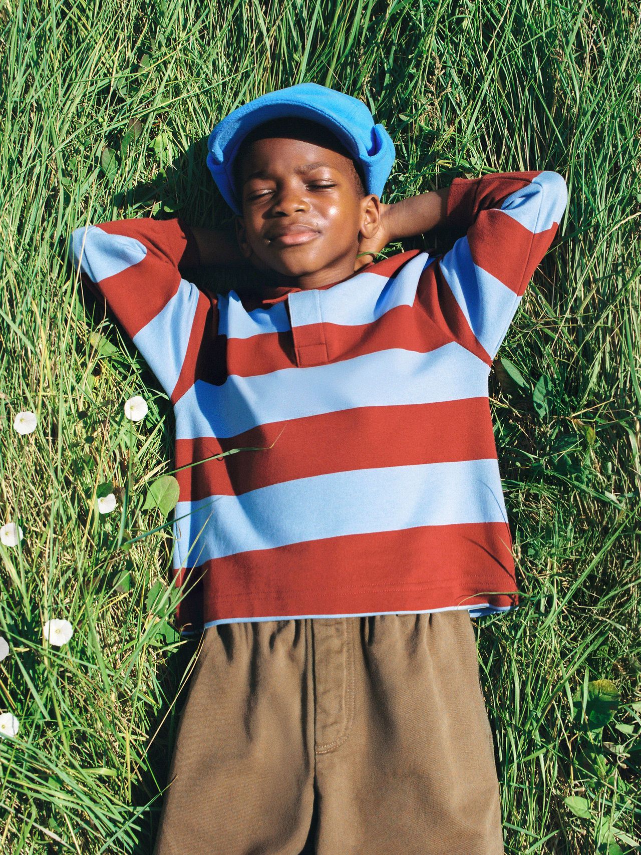 Child in a striped shirt and blue cap lies on grass with eyes closed, arms behind head, surrounded by small white flowers.