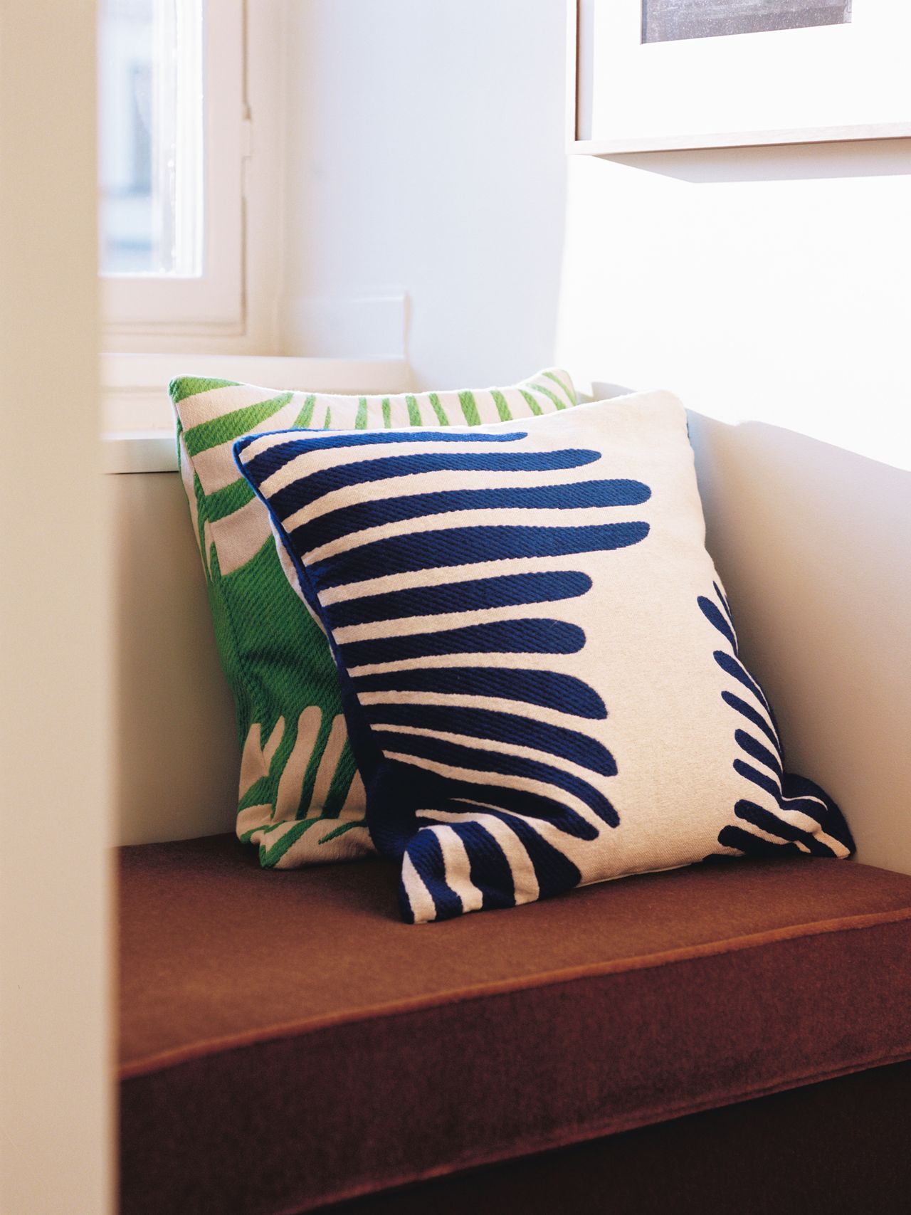 Two decorative pillows with abstract blue and green patterns on a brown cushioned bench by a sunlit window.