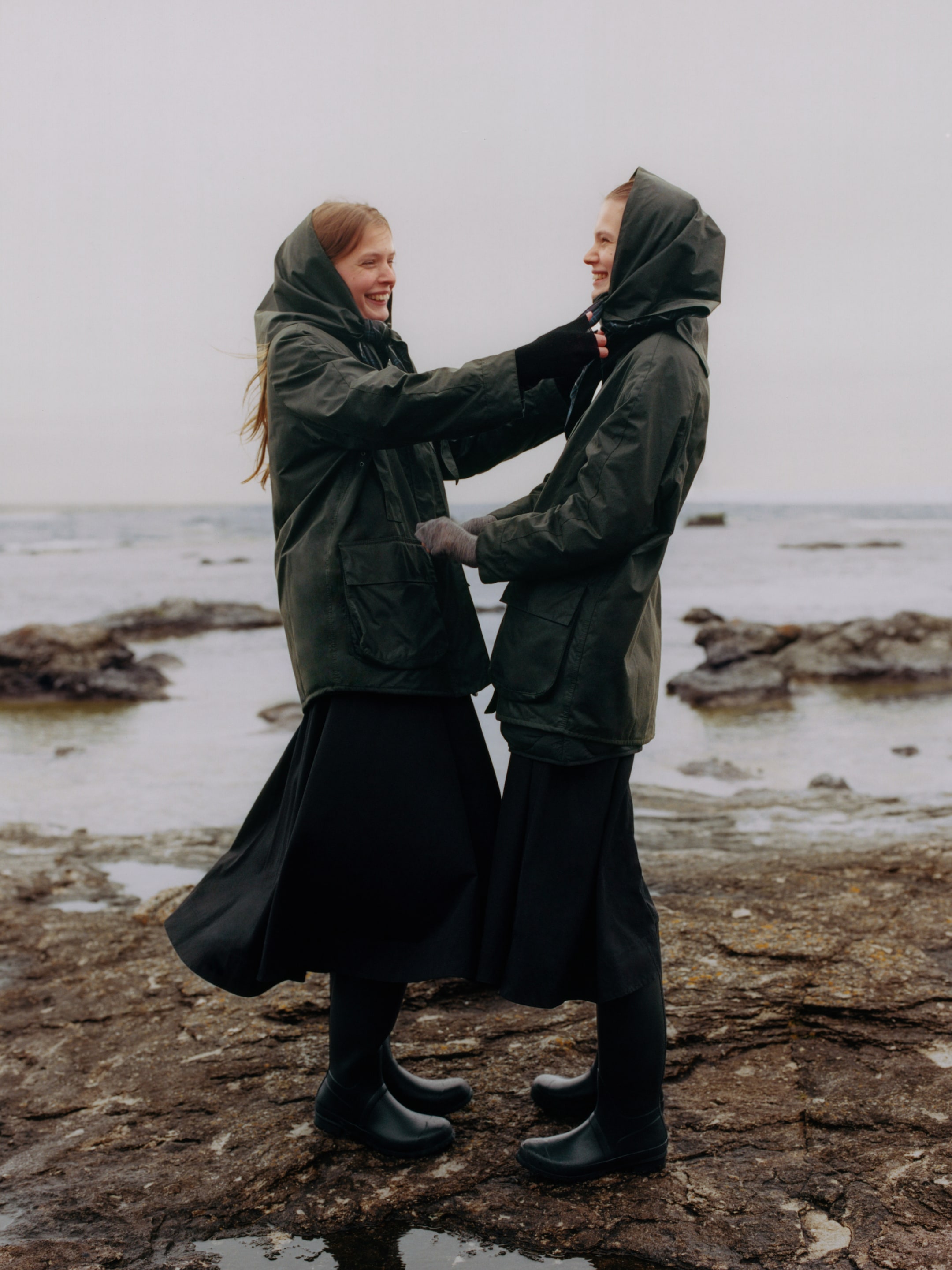 Two people in dark coats and boots stand on a rocky shore, smiling at each other. The ocean is visible in the background.