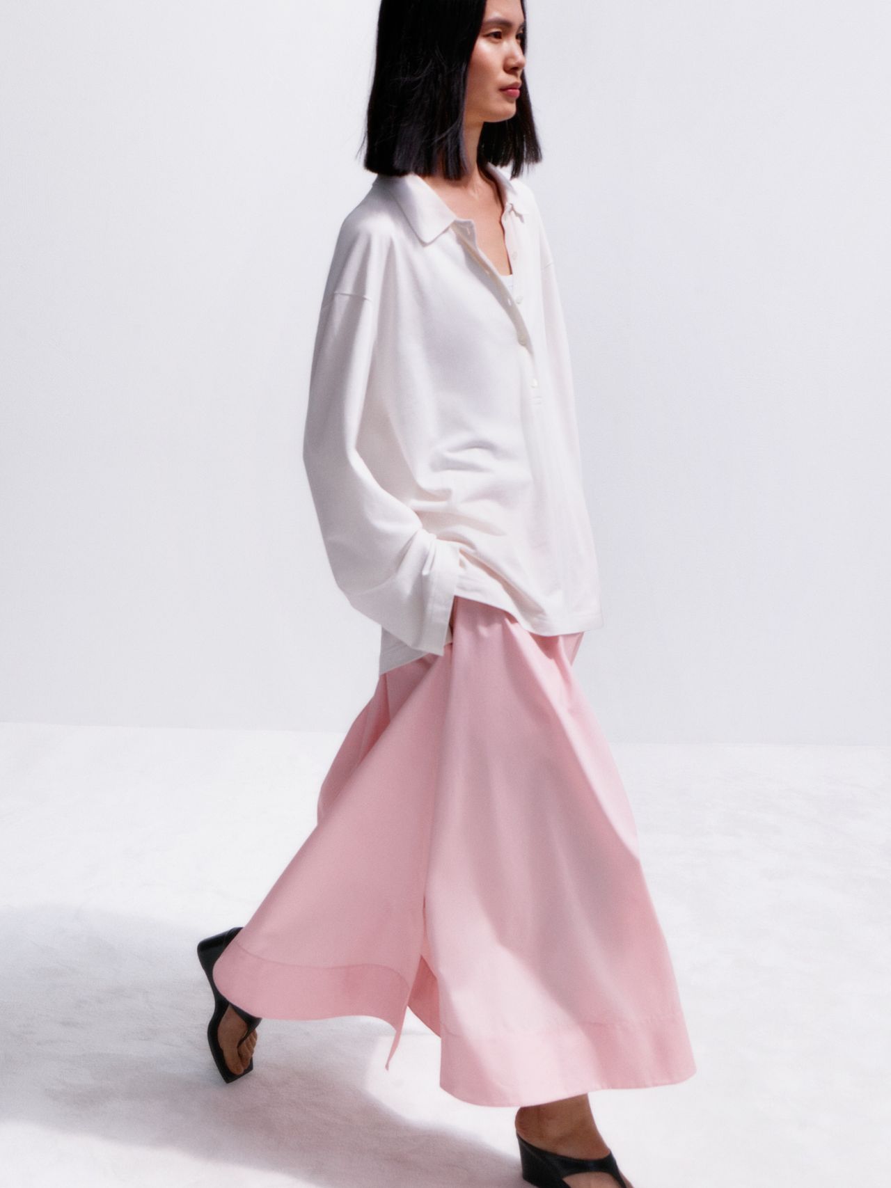 Woman in a white blouse and flowing pink skirt walking against a light background, wearing black heels.