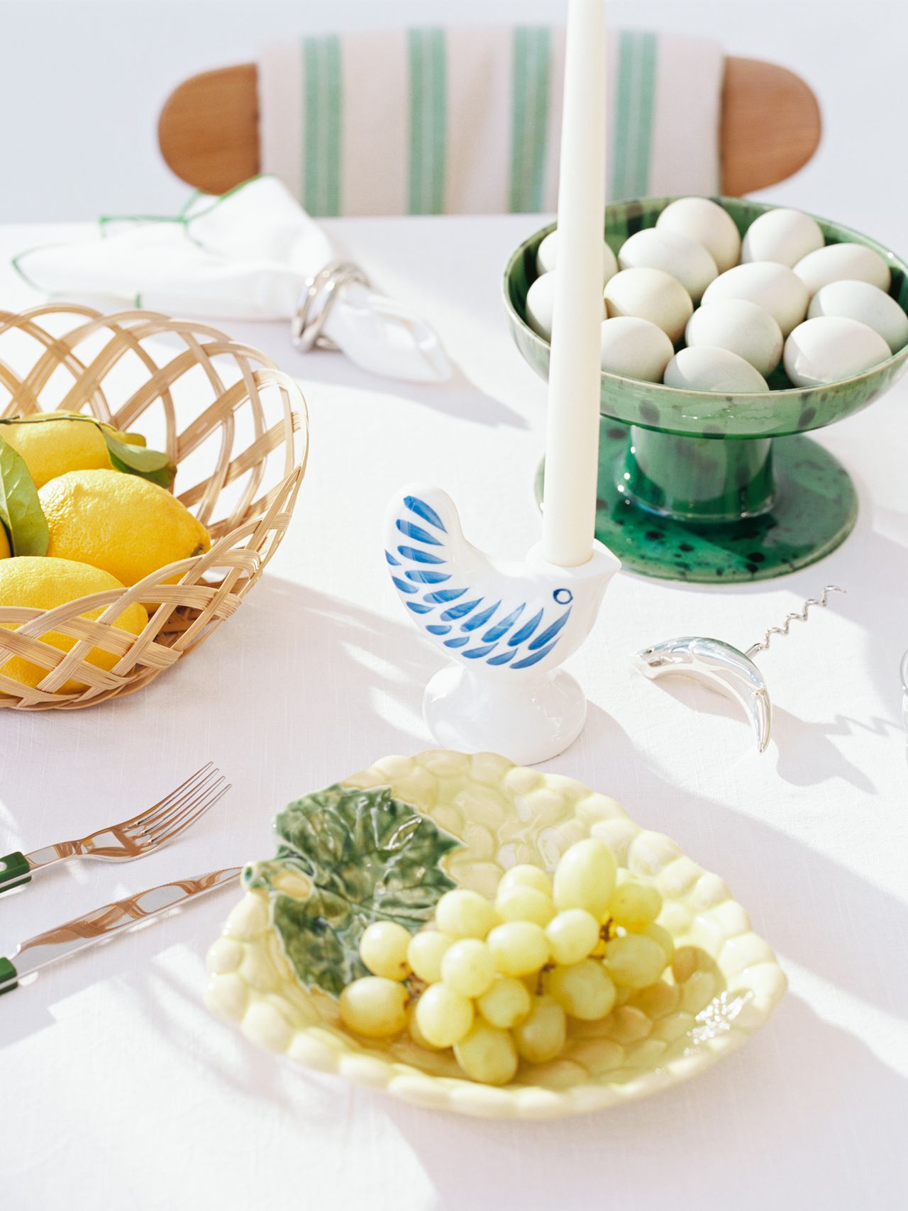 A table setting with a basket of lemons, grapes on a leaf-shaped plate, a candle in a bird holder, and a bowl of white eggs.