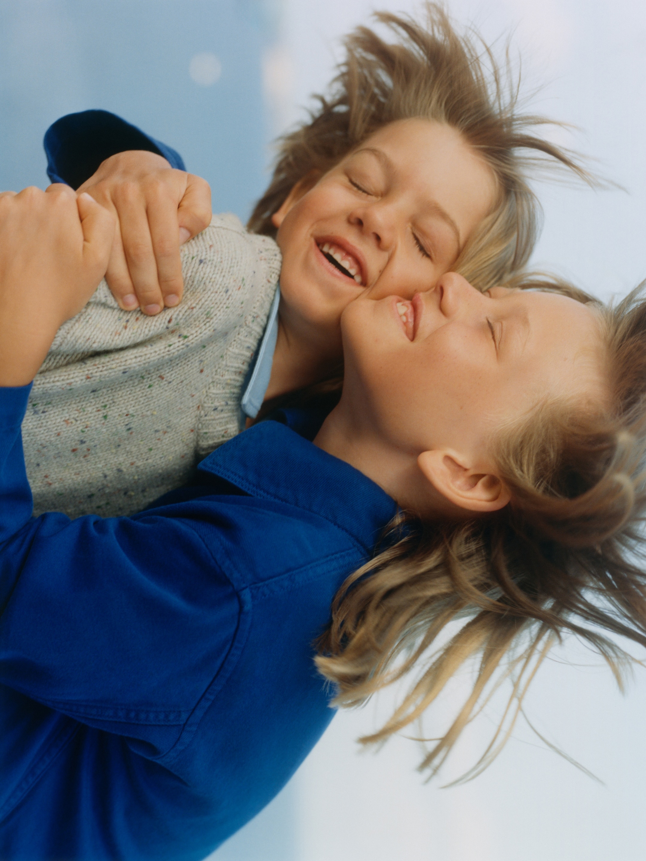 Two children with eyes closed and hair blowing in the wind share a joyful embrace, wearing casual clothes against a blurred, light background.