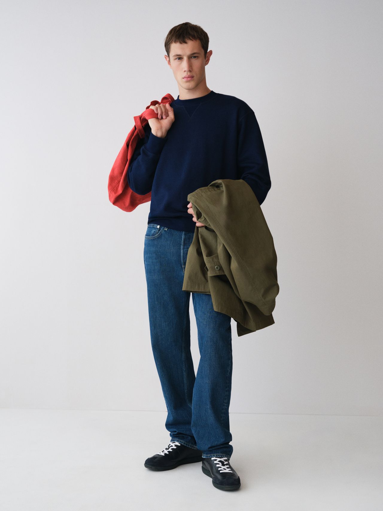 Person in a navy sweater and jeans holds a red and an olive-green jacket, standing against a plain white background.