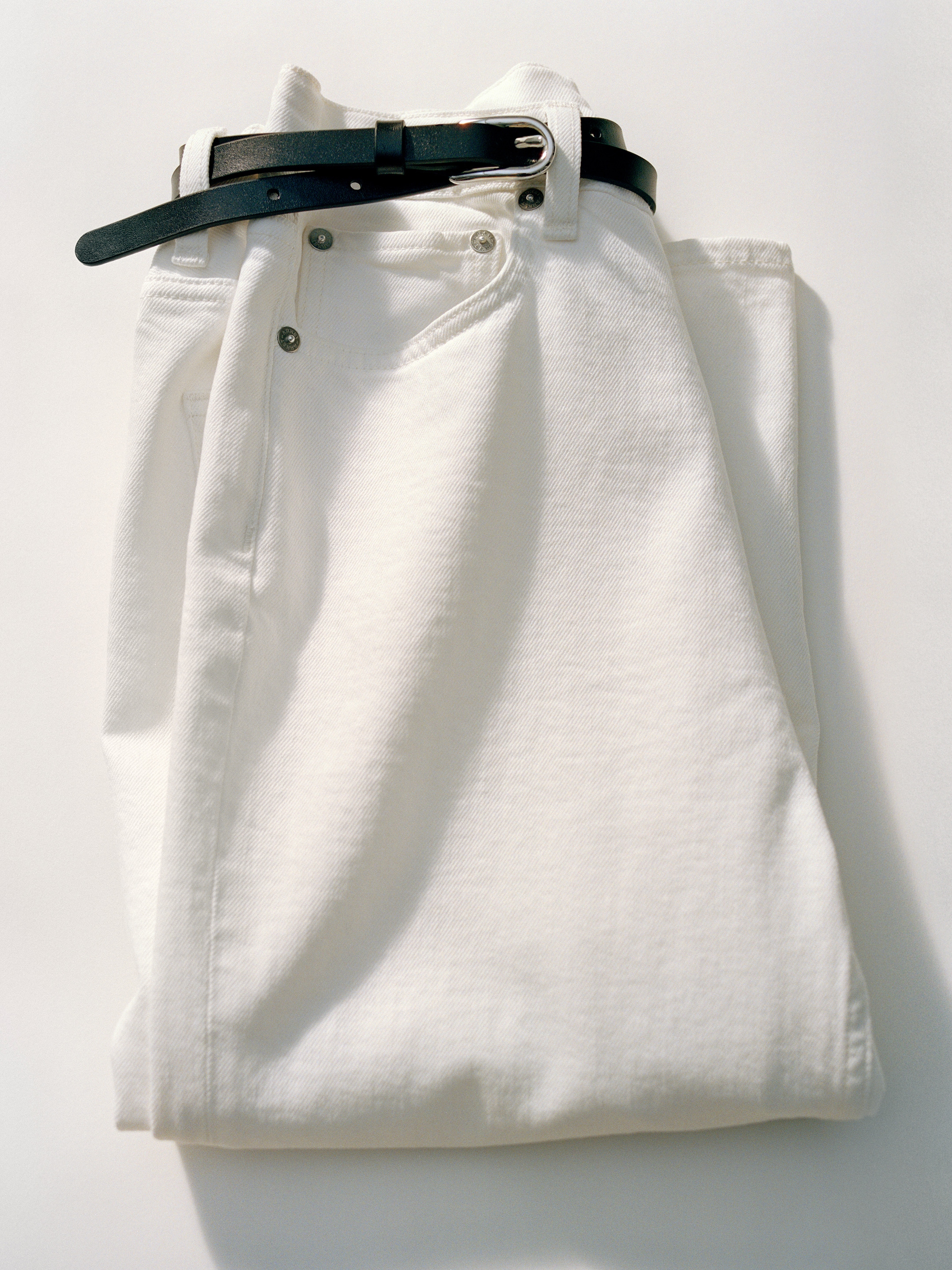 Folded white jeans with a black leather belt threaded through the loops, placed on a light surface.