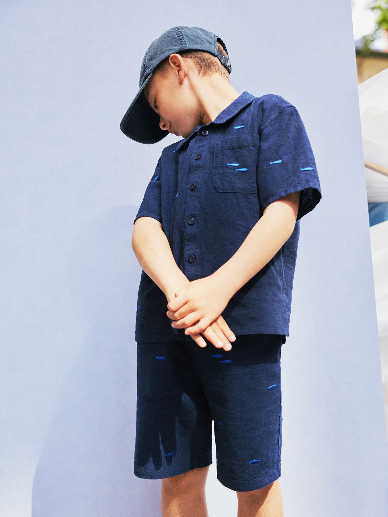 Child in blue shirt and shorts with subtle patterns, wearing a matching cap, stands against a light blue background, looking down.