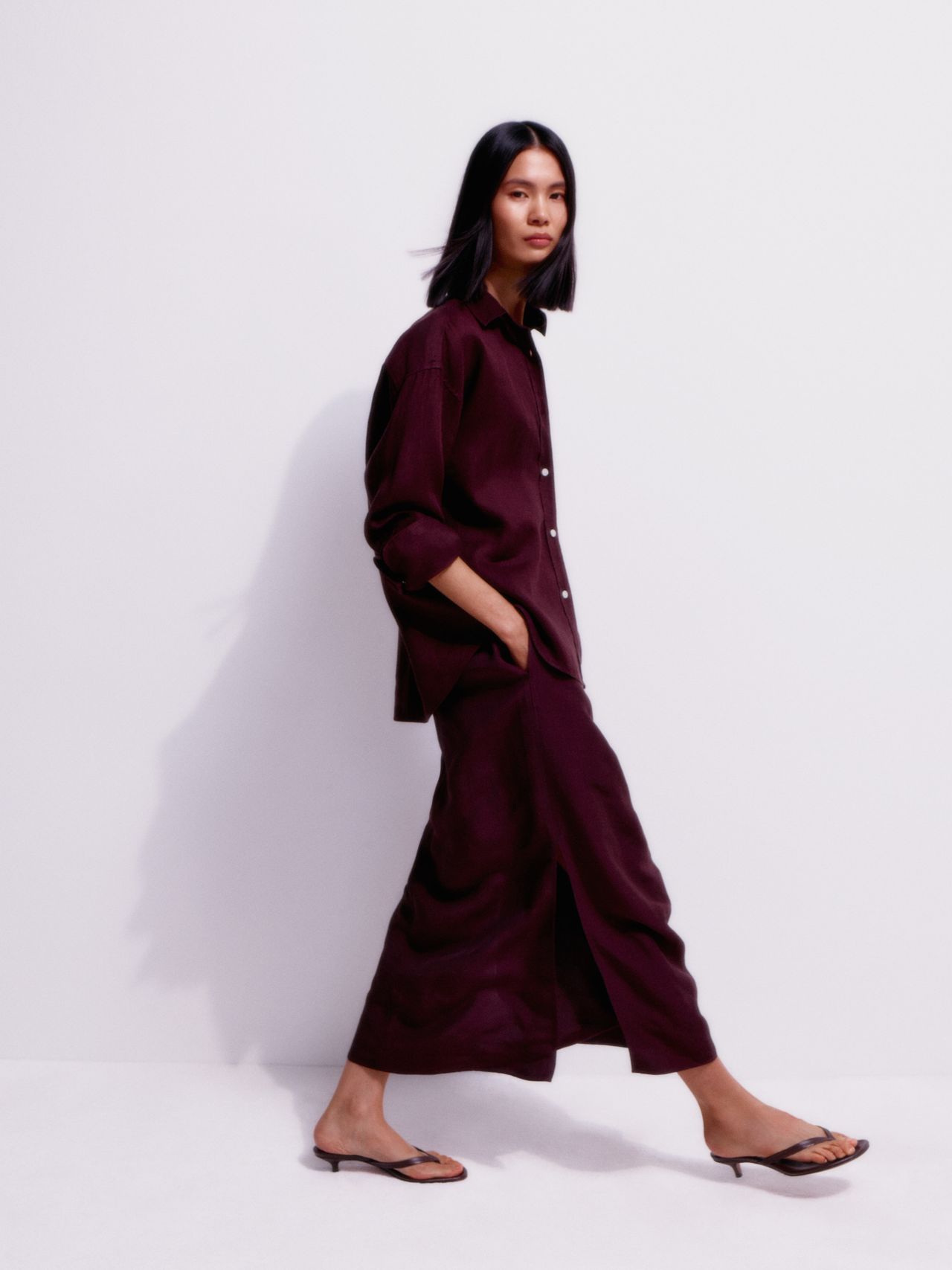 Woman in a burgundy shirt and skirt walks against a white background, wearing flip-flops and with hands in pockets.