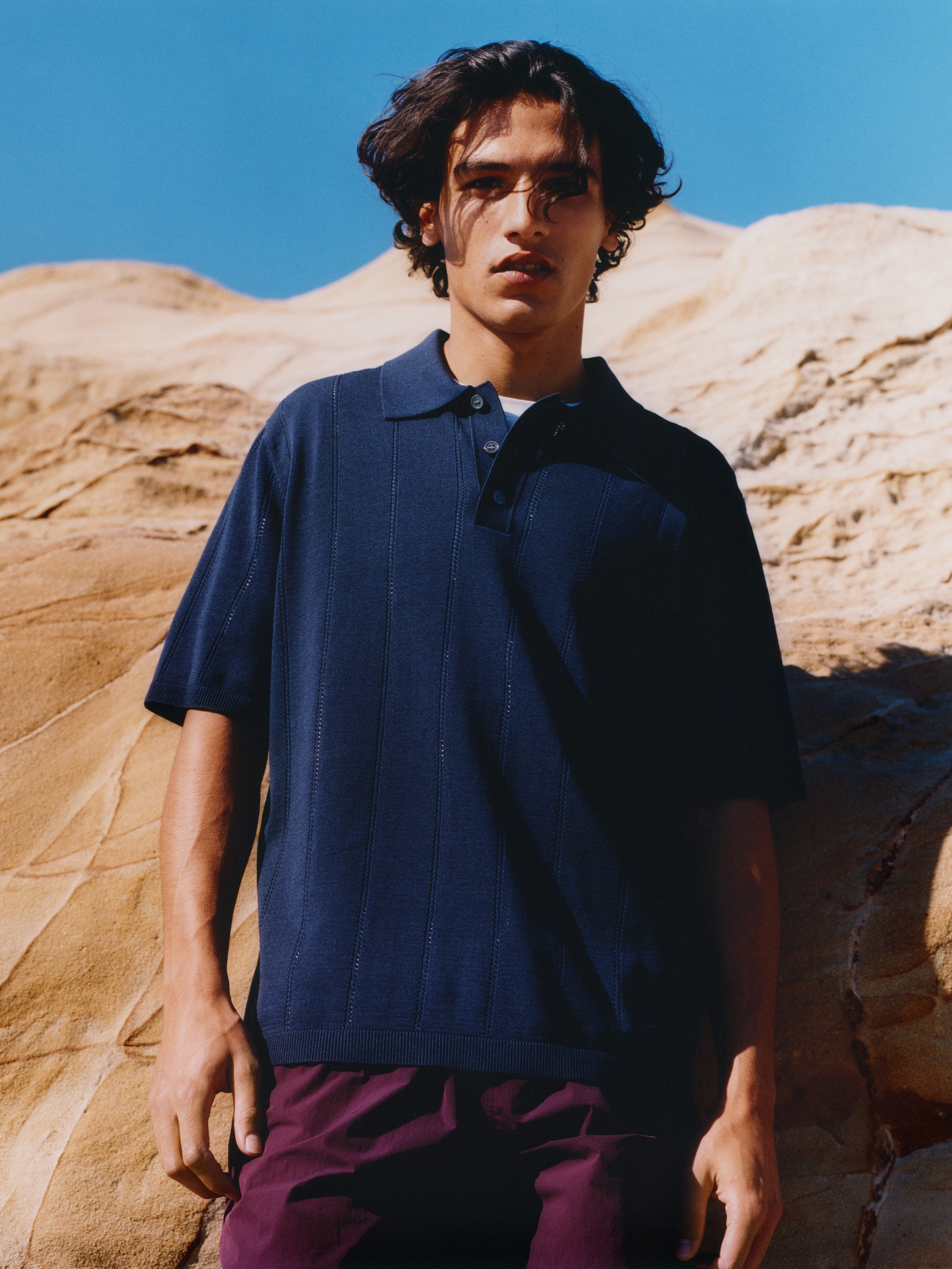 Young man in a navy polo shirt and maroon pants stands against a backdrop of sandy rock formations under a clear blue sky.