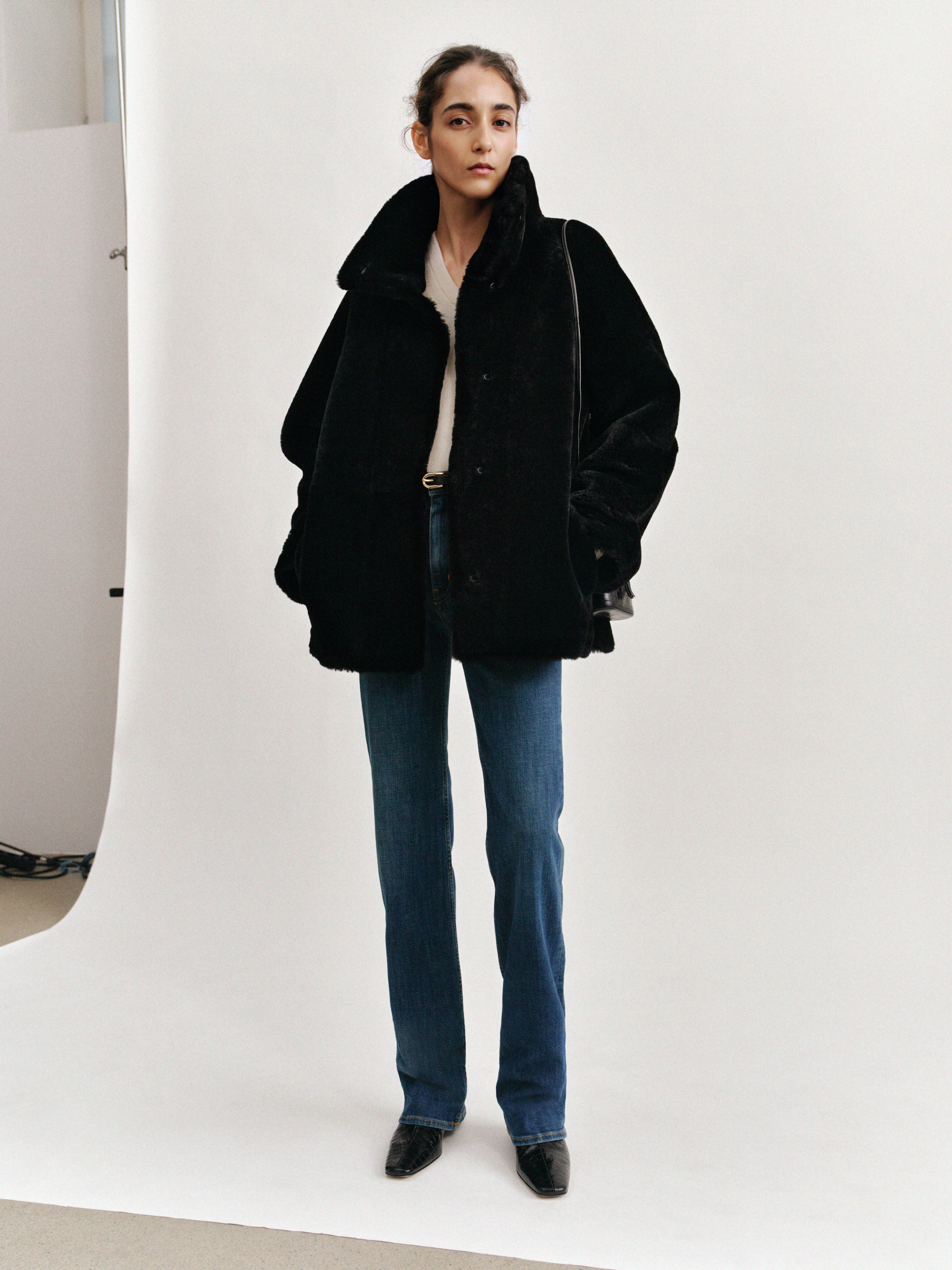 A person wearing a black coat, white top, blue jeans, and black shoes stands against a plain white background, looking at the camera.
