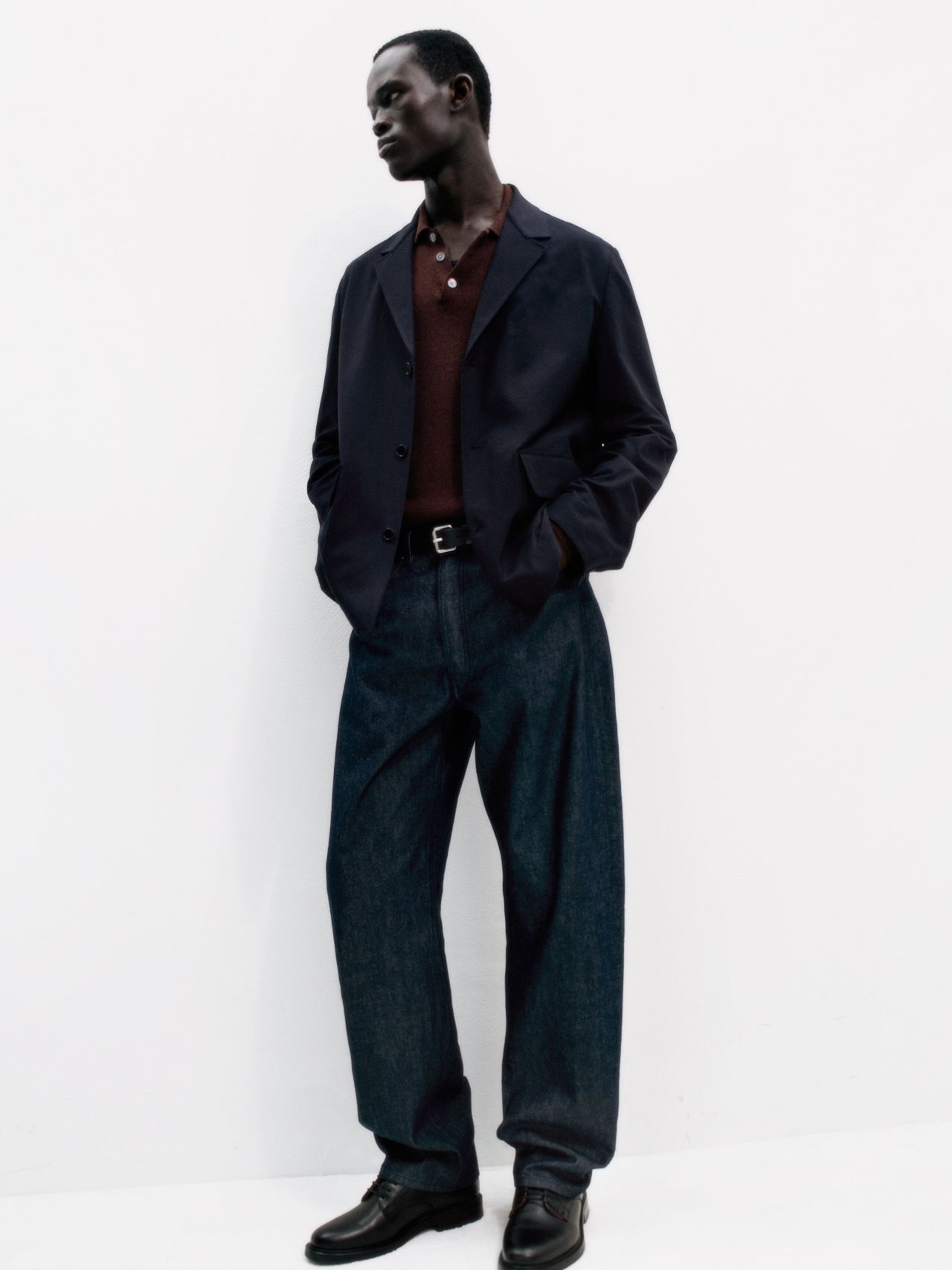 Man in a navy blazer, maroon shirt, and loose-fit jeans standing against a white background, hands in pockets, looking left.